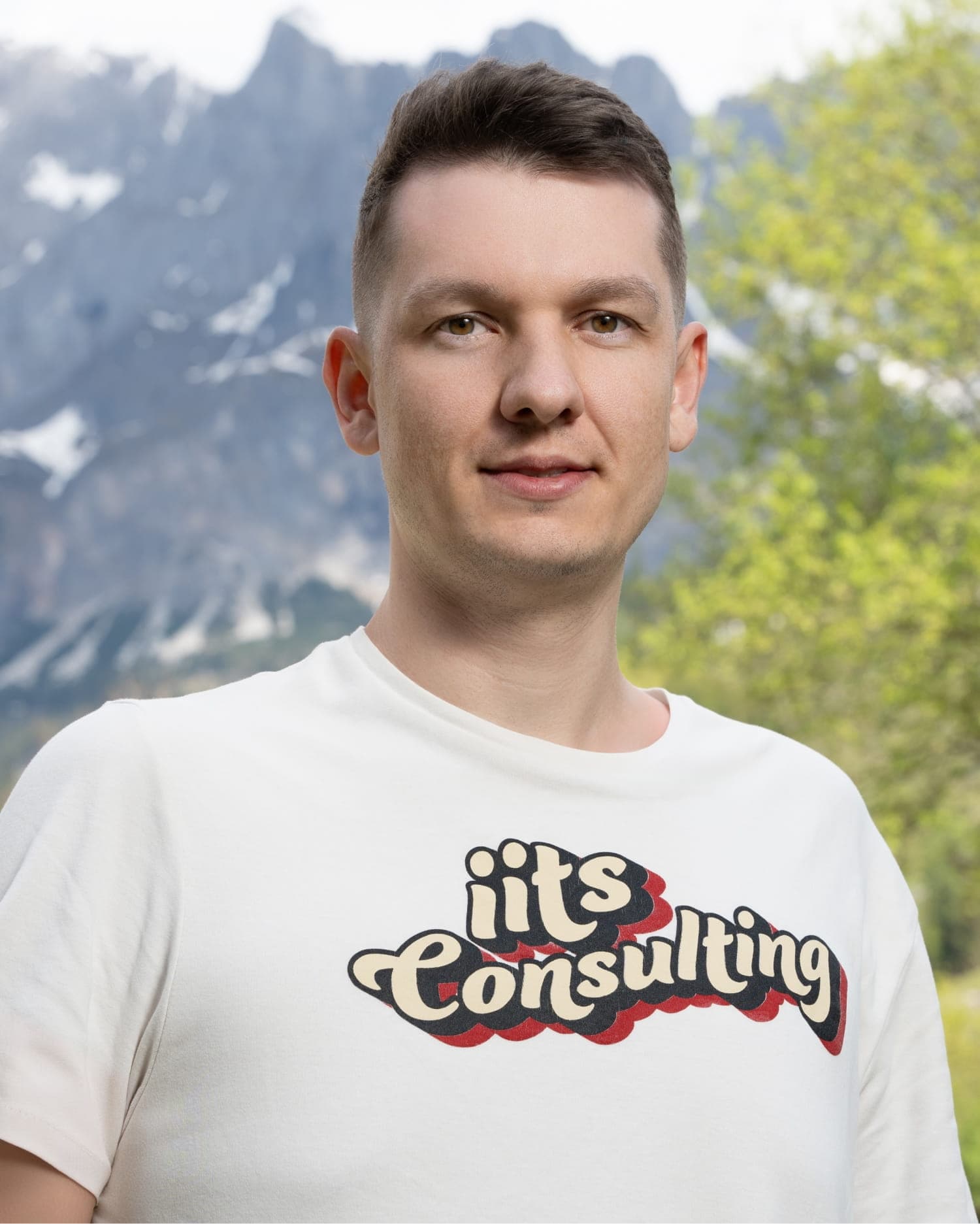 Portrait of Artem Lajko, a young man with short brown hair and light brown eyes, wearing a plain white t-shirt with an 'iits Consulting' logo in a retro-style cream and red font. He is smiling outdoors with a snow-dusted mountain and green trees in the background.