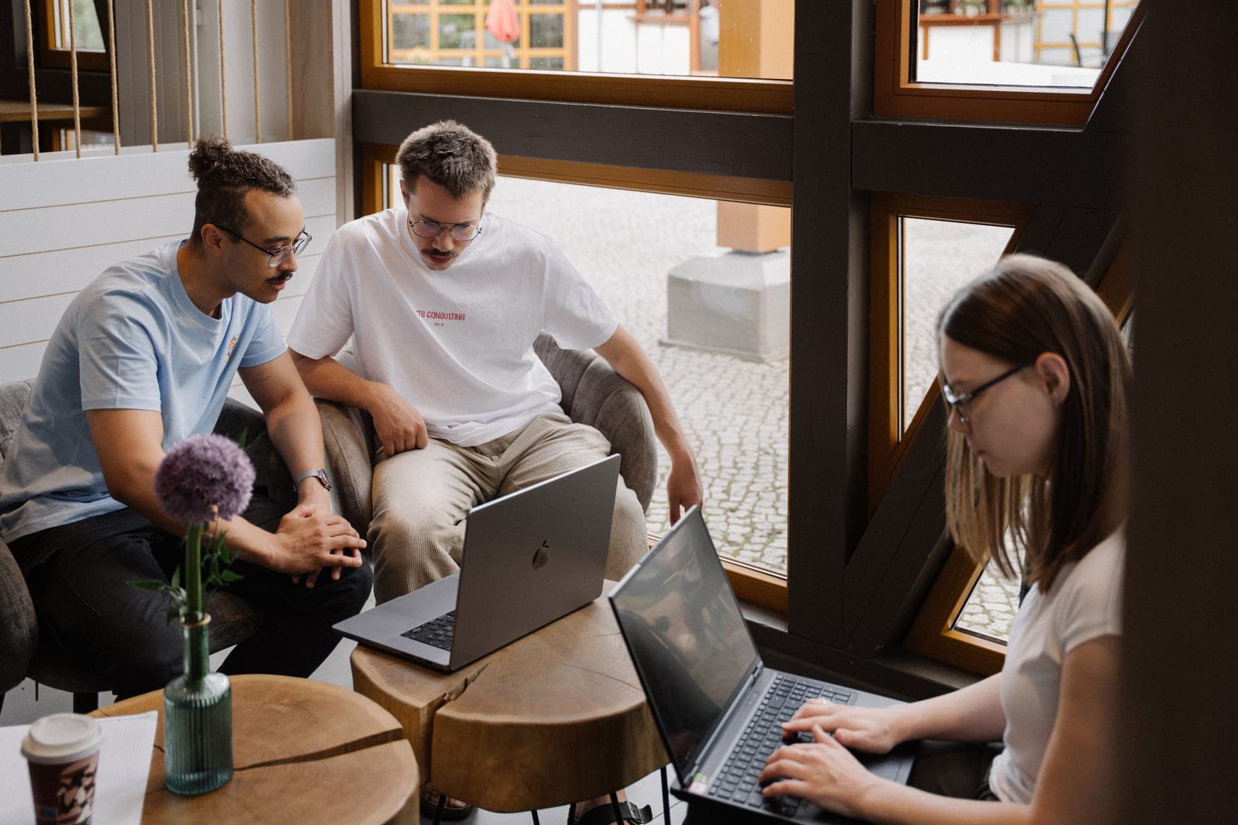 Two men and a woman collaborating in a modern workspace, focused on a laptop screen near a rustic table and a purple flower.