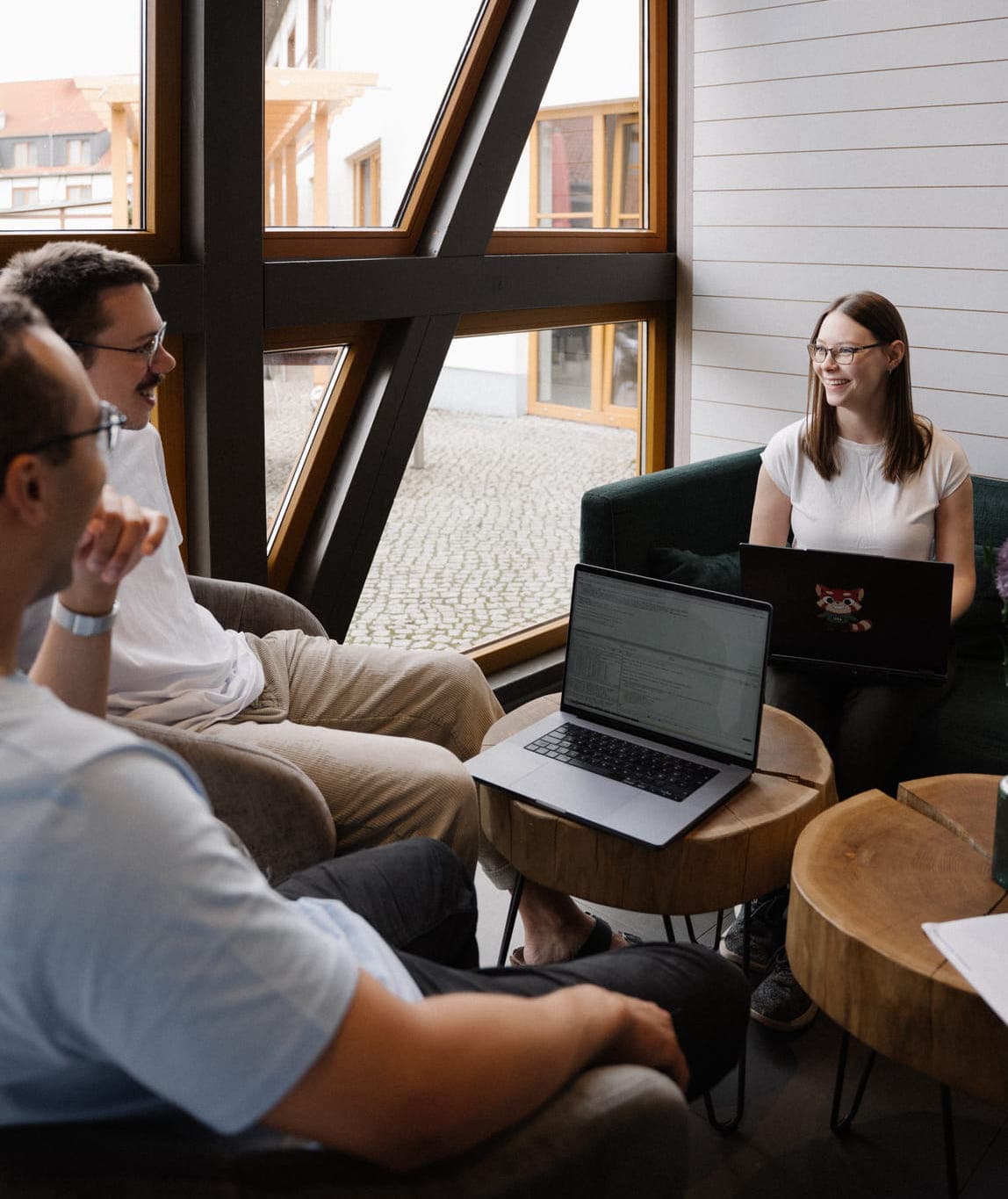 A small group sits in a lounge with laptops, chatting and working together near large windows.