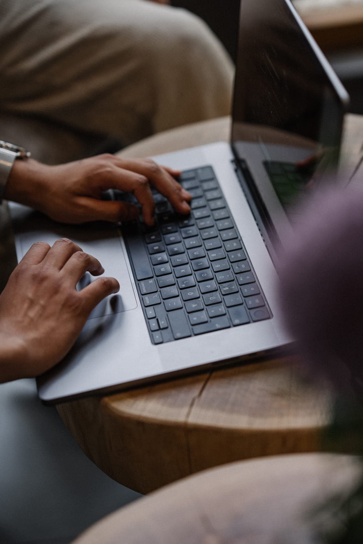 A close-up shot of a person's hands typing on a silver laptop, which is resting on a round wooden table.