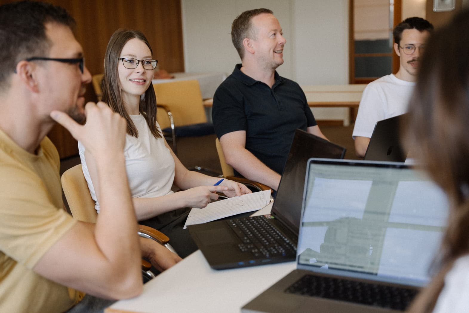 A group of colleagues sitting around a table with laptops, smiling and looking off-camera to the right.