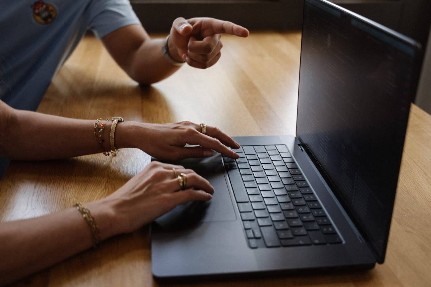 Hands type on a laptop at a wooden table while someone nearby points at the screen.