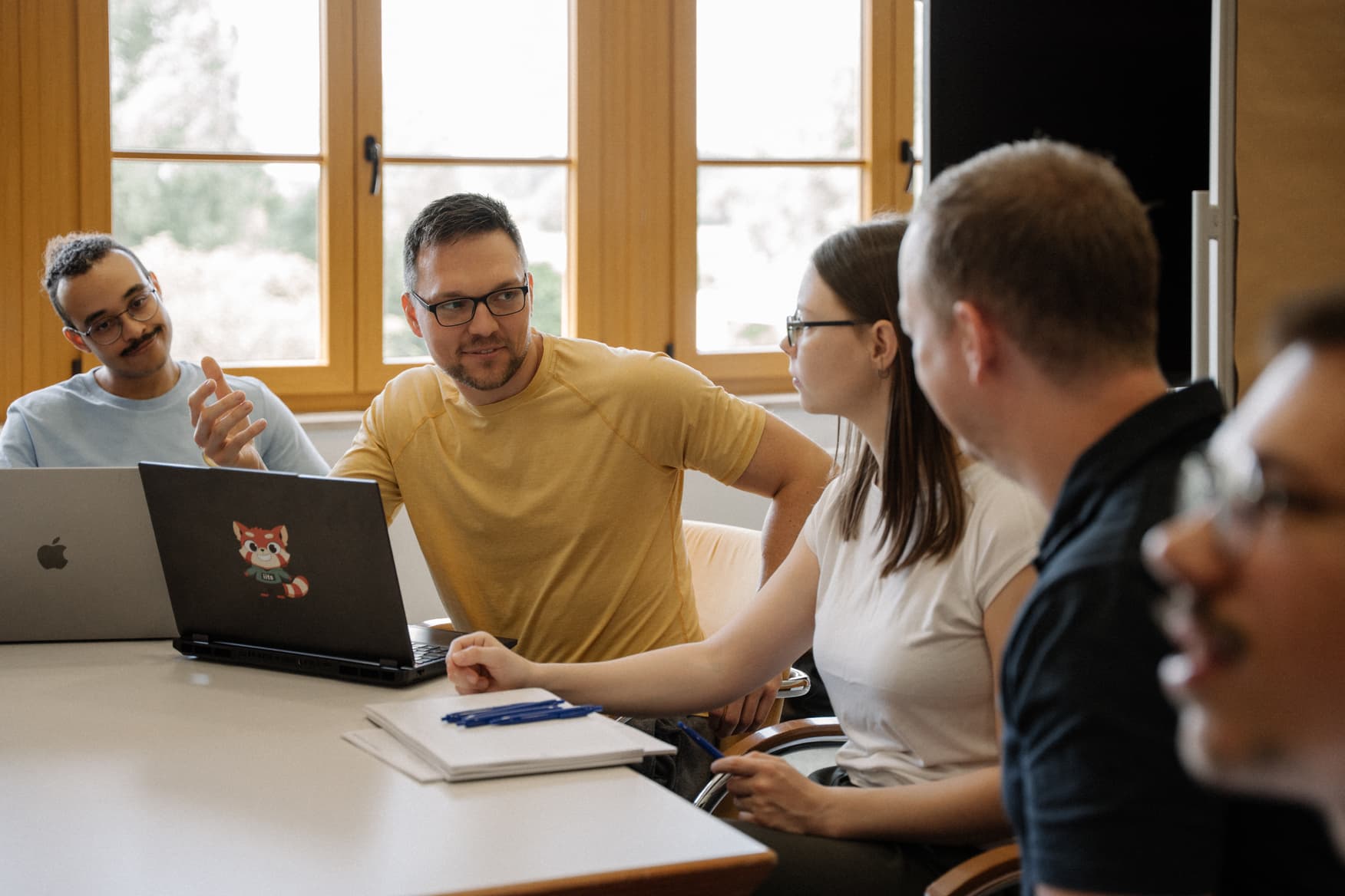 Five people seated around a white table engaged in a collaborative meeting; a man in a yellow t-shirt gestures while speaking to a woman near a laptop with a red panda sticker, while another man in a blue shirt smiles nearby.