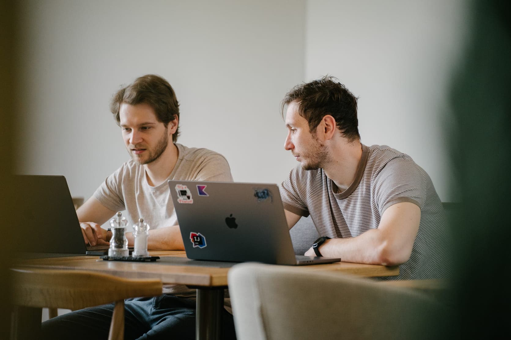 Two colleagues sitting at a wooden table, each focused on their own laptop.