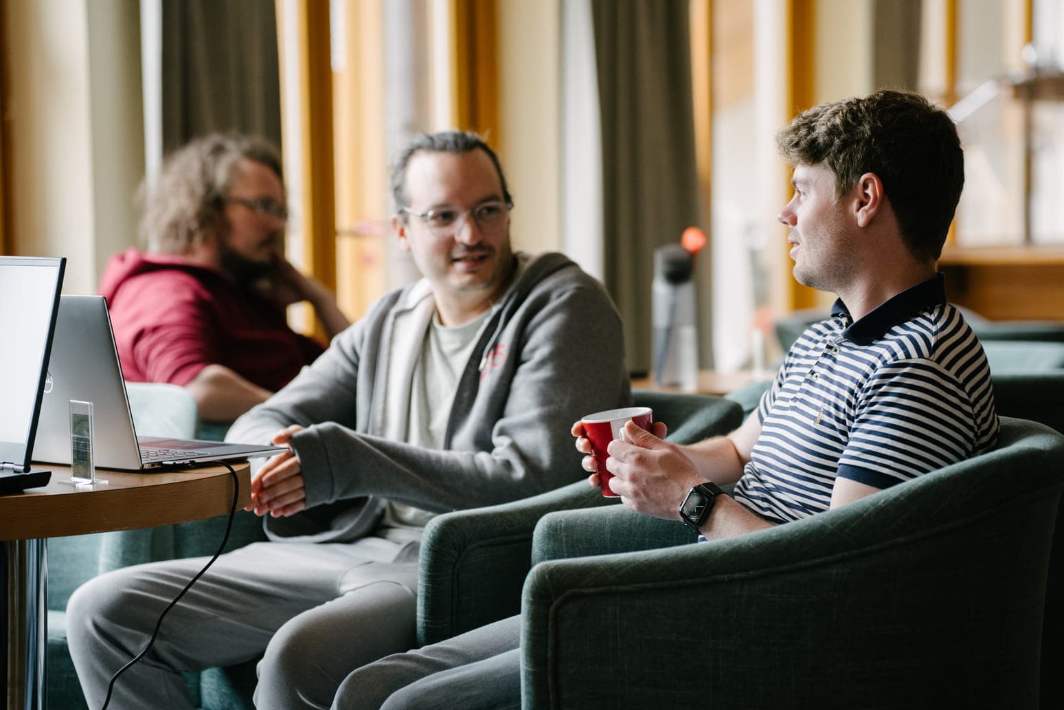 Three people sit on green armchairs in a bright lounge, chatting beside a laptop and a small table.