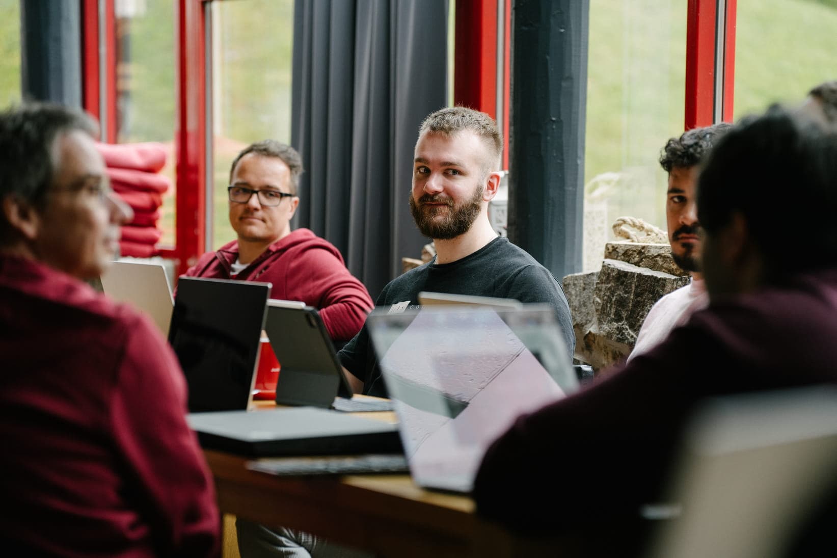 A group of men sitting around a table with laptops, participating in a meeting or workshop. One man with a beard and a black t-shirt smiles at the camera, while others look on.