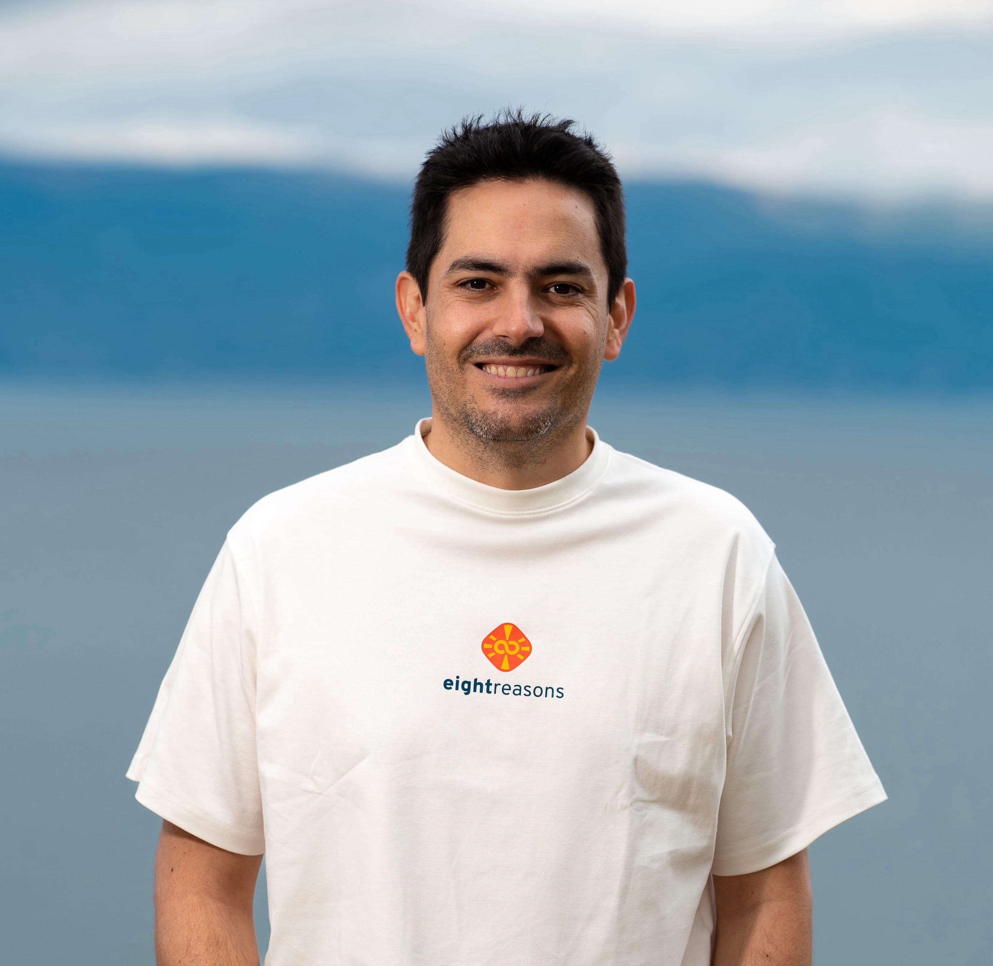 Pablo Uribe, smiling with short dark hair and light stubble, wearing a white eightreasons t-shirt, standing against a backdrop of water and sky..