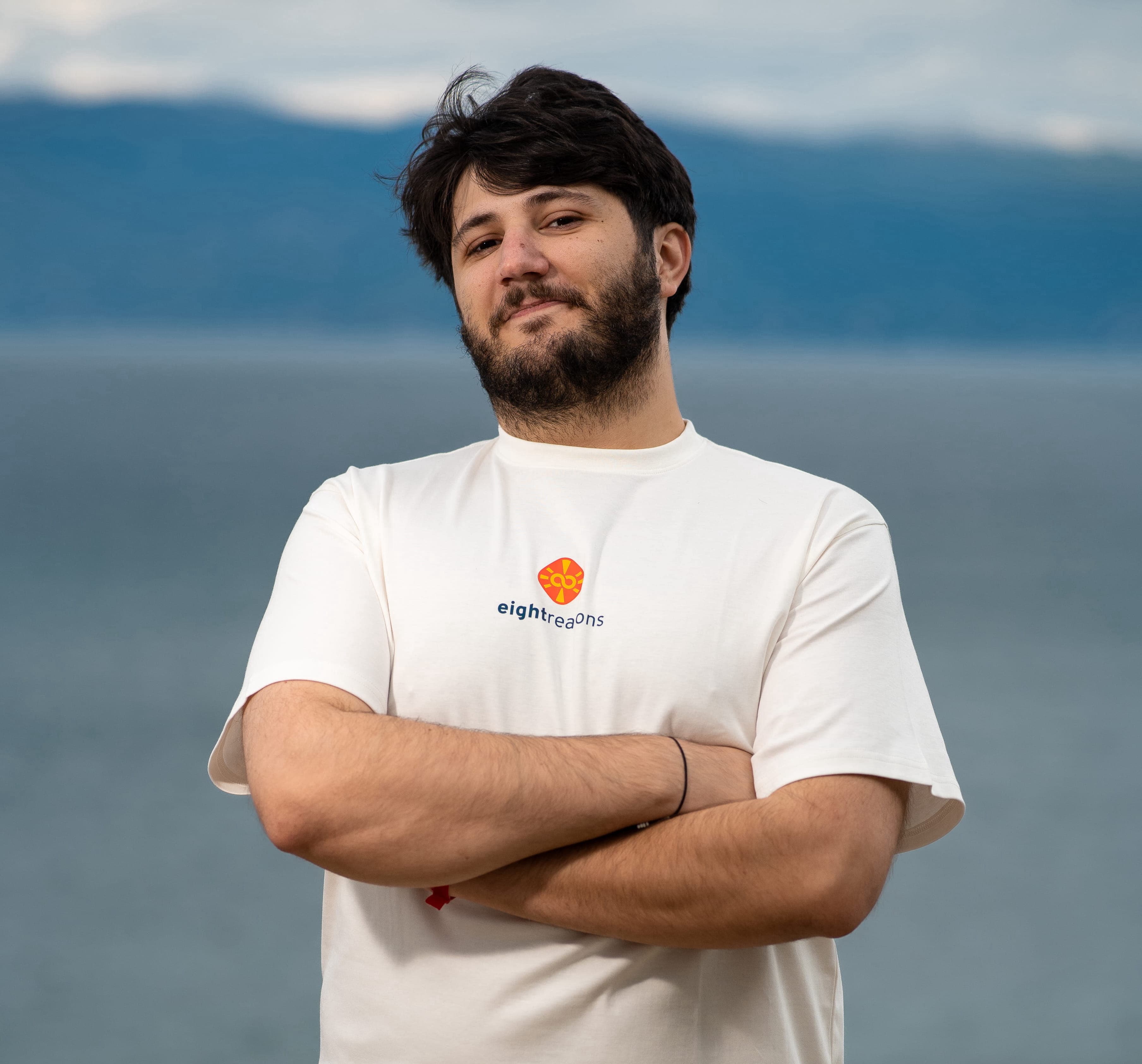 Alija Nuredini, with dark hair and a beard standing with his arms crossed, wearing a white eightreasons t-shirt, against a water and sky background.