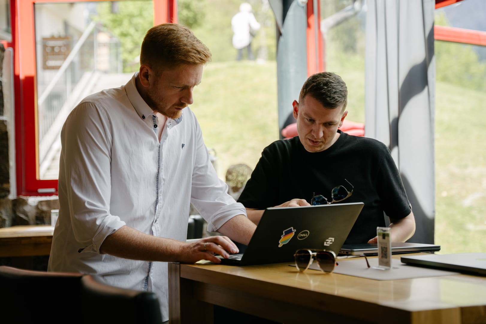 Two colleagues working together at a wooden table near a window, focusing on a laptop while a person walks by outside.