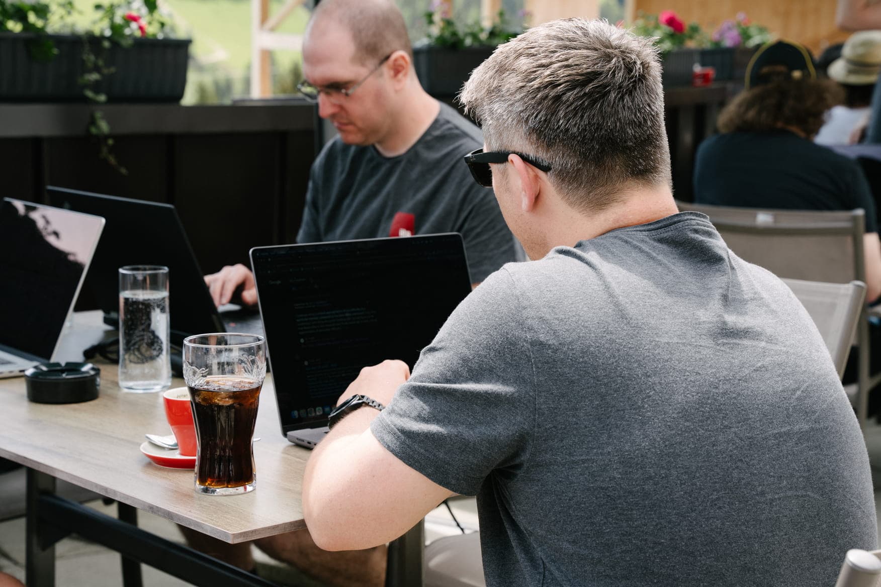 Three people work on laptops at an outdoor table, with drinks and a relaxed café setting around them.