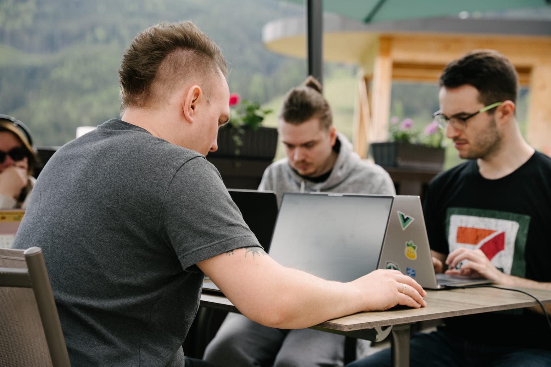 Three colleagues working on laptops at a table on an outdoor terrace, with green hills visible in the background.
