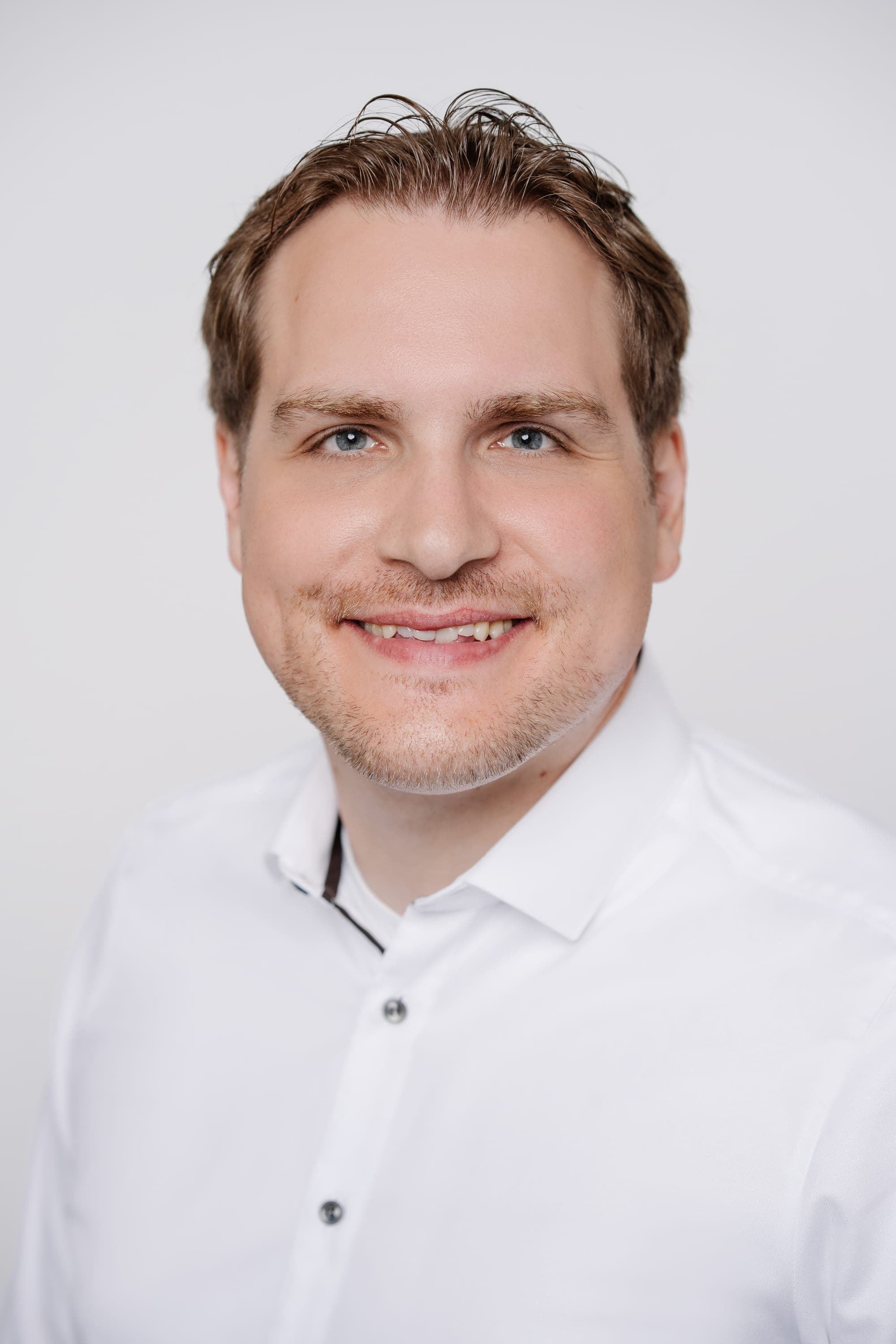 Portrait of Dr. Tim Delbrügger, a man with short light brown hair and blue eyes, wearing a white button-down shirt with the top button undone. He is smiling gently against a plain white background