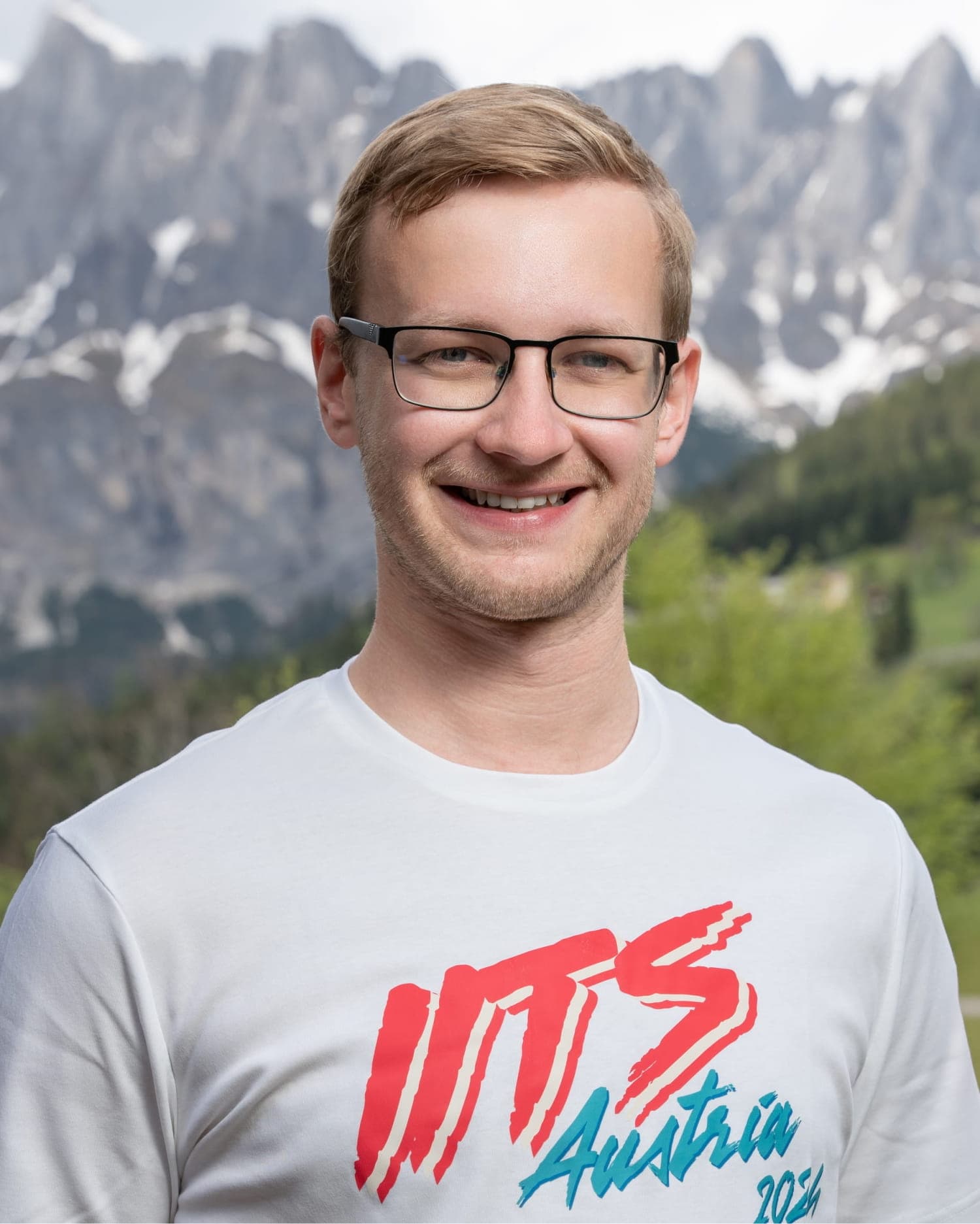 Portrait of Christoph Aufmhoff, a man with short light brown hair and rectangular glasses with thin black frames, wearing a white t-shirt with an 'ITS Austria 2024' graphic. He is standing outdoors with a green, grassy landscape and snow-capped mountains in the background.