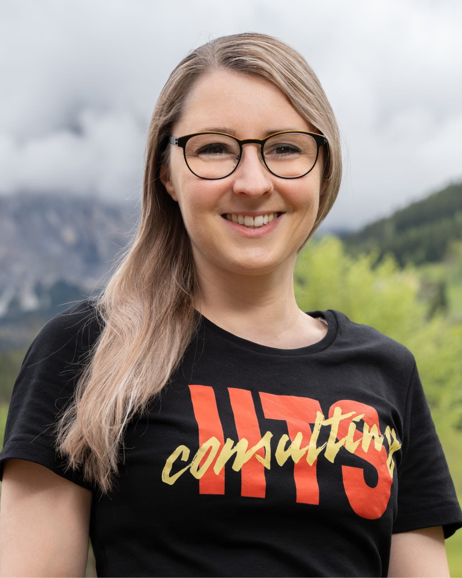 Portrait of Anna Karina Flister, a woman with long, straight blonde hair, wearing a black t-shirt with a red and gold 'iits consulting' logo. She is smiling warmly outdoors with green hills and mountains in the background.
