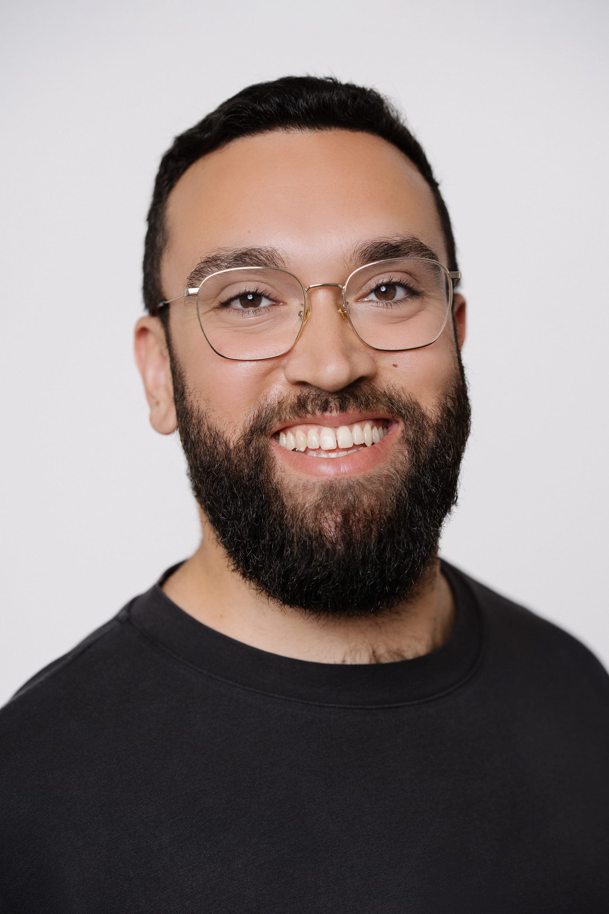 Portrait of Маx Knopf, a man with short dark hair, a full beard, and thin gold-rimmed glasses. He is smiling at the camera and wearing a black crew-neck t-shirt against a plain light gray background