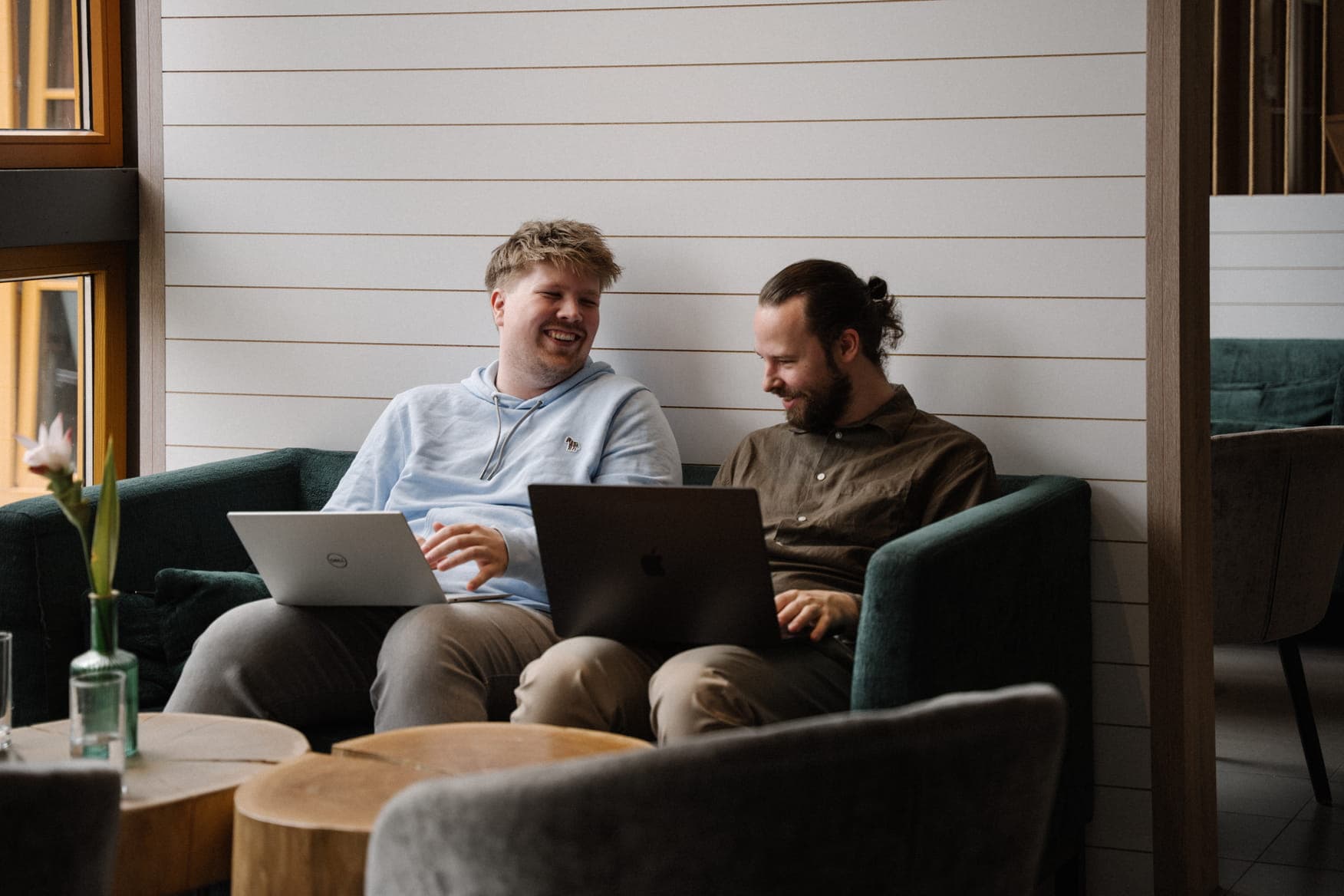 Two men sitting side-by-side on a dark green sofa, both using laptops. The man on the left wears a light blue hoodie and is smiling broadly, while the man on the right, with long hair and a beard, wears a brown shirt. They are in a room with white horizontal wall paneling.