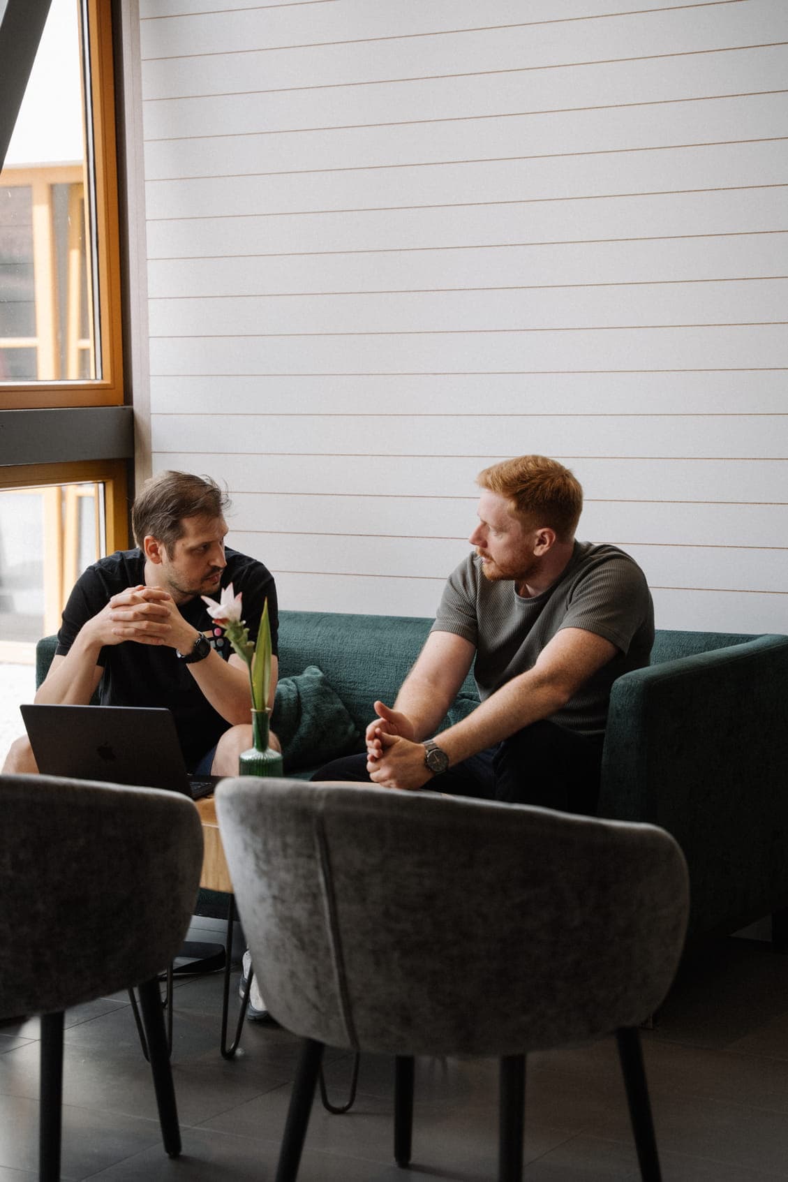 Two men seated on a teal sofa engaged in a conversation. The man on the left wears a black t-shirt with a laptop on his lap, while the man on the right, with red hair and a beard, wears a grey t-shirt. A small wooden table with a green vase holding a white flower sits in front of them.