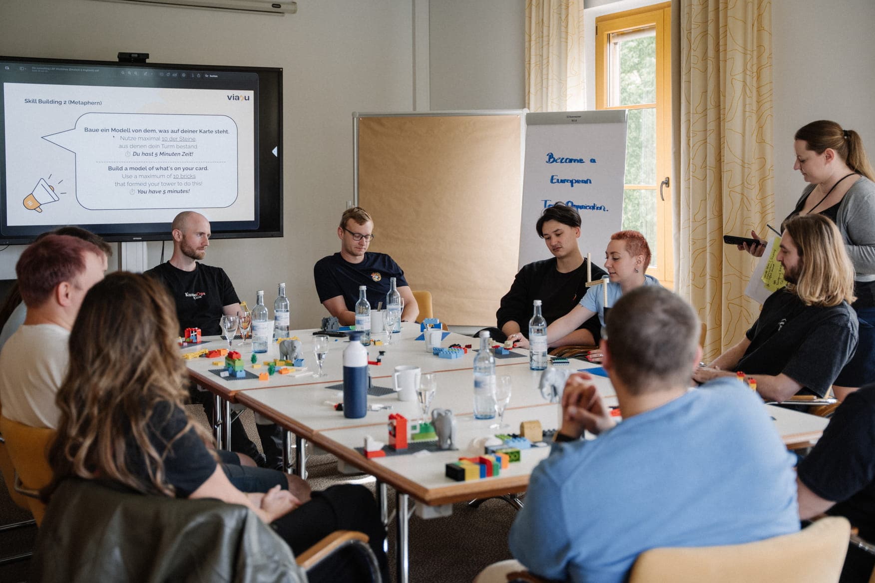 A corporate workshop in progress where participants build models from colorful blocks, guided by a presentation slide titled 'Skill Building 2 (Metaphern)'.