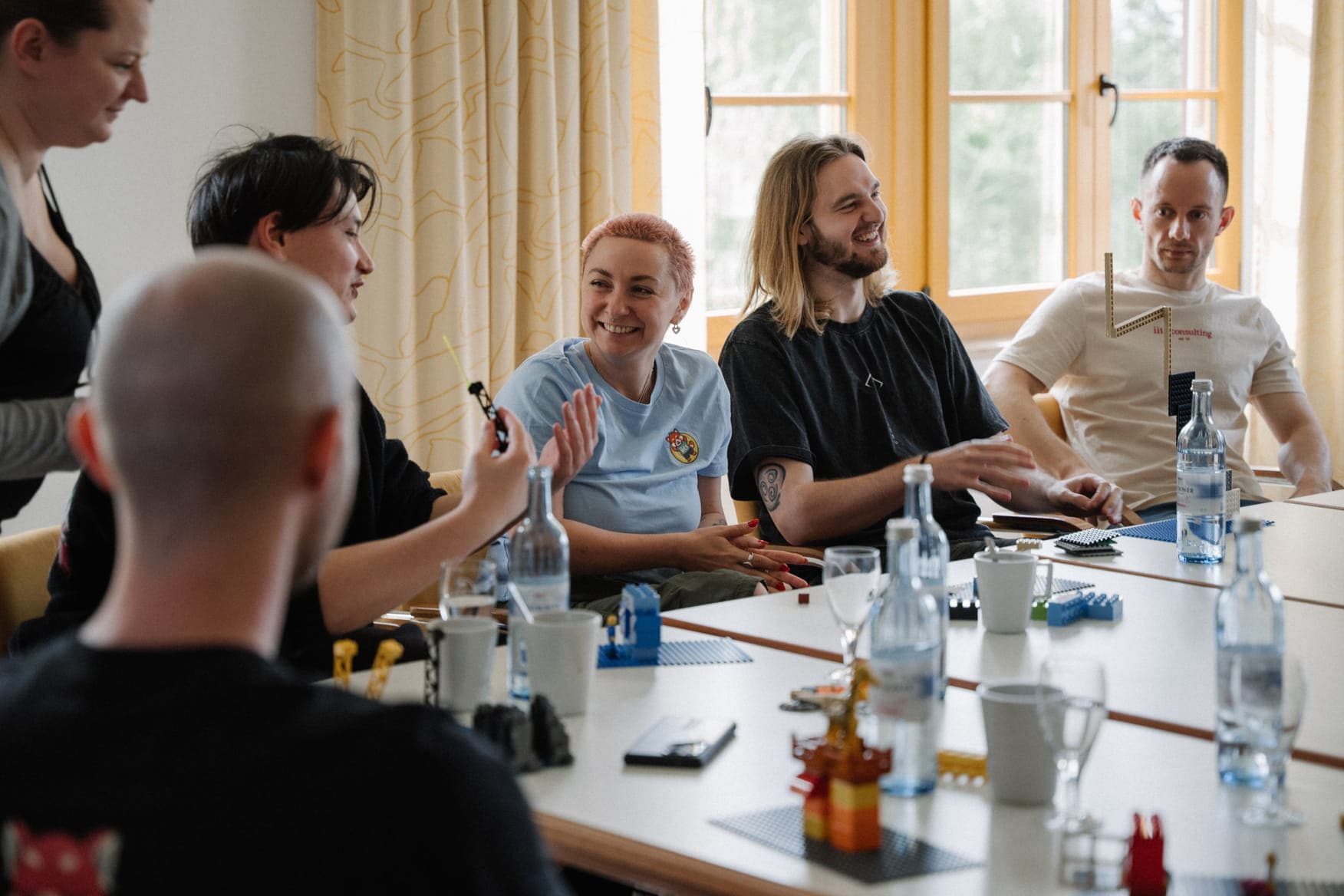 A group of people sitting around a long table in a bright room, laughing and interacting during a workshop. The table is scattered with water bottles, mugs, and colorful LEGO models. A woman with short pink hair smiles while clapping.