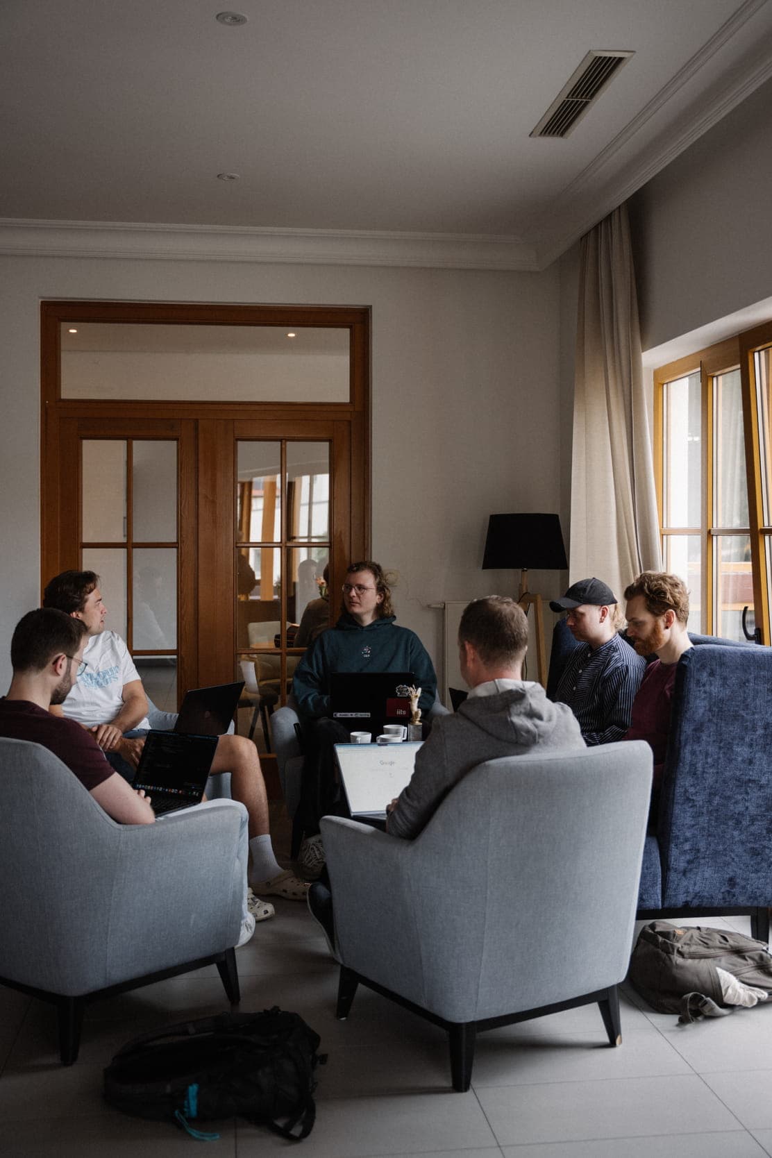 A group of six colleagues working collaboratively on laptops while seated in a casual circle of armchairs and a sofa in a bright room.