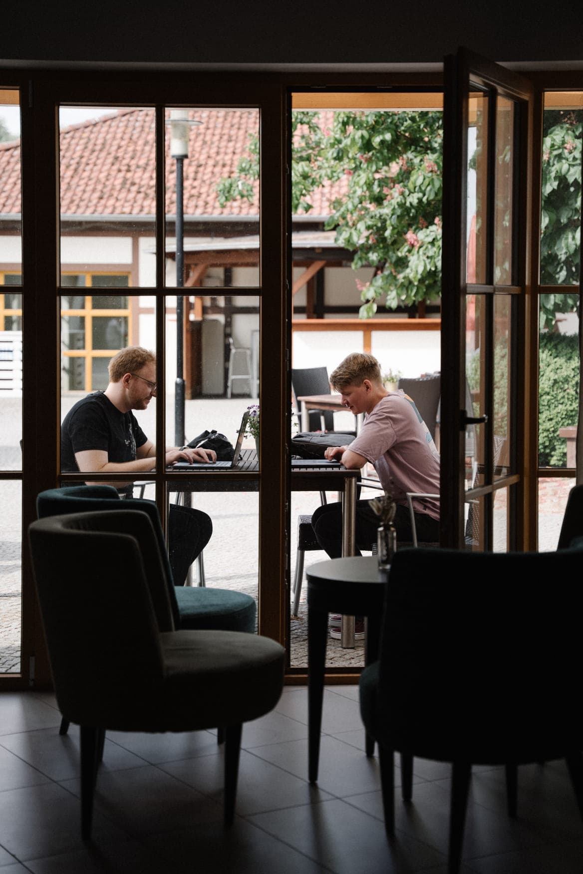 Two people sitting at an outdoor table working on laptops, viewed through large, open wooden doors from a dimly lit interior.