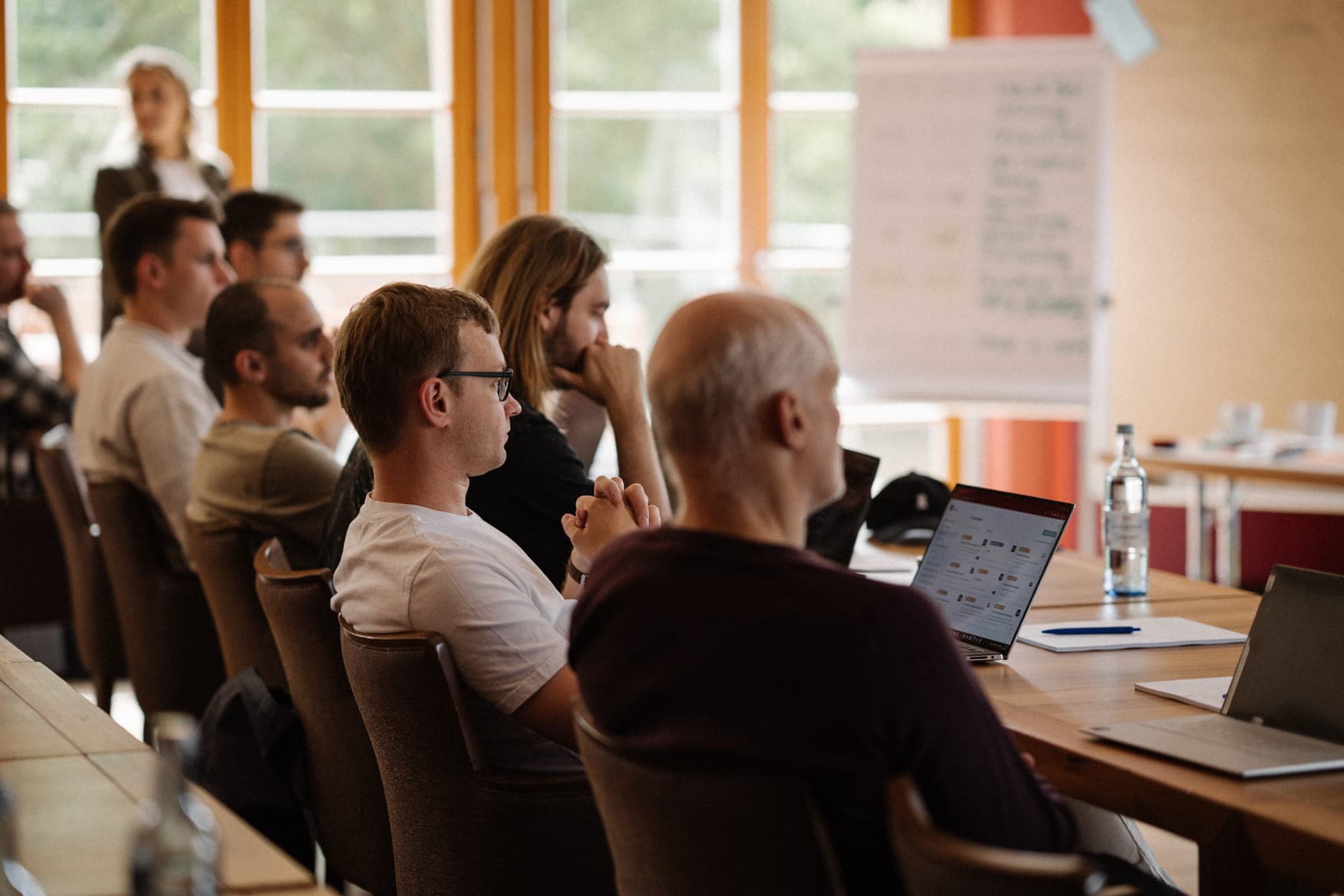 Team members seated at long wooden tables in a well-lit room, listening attentively to a presentation with open laptops and notebooks.