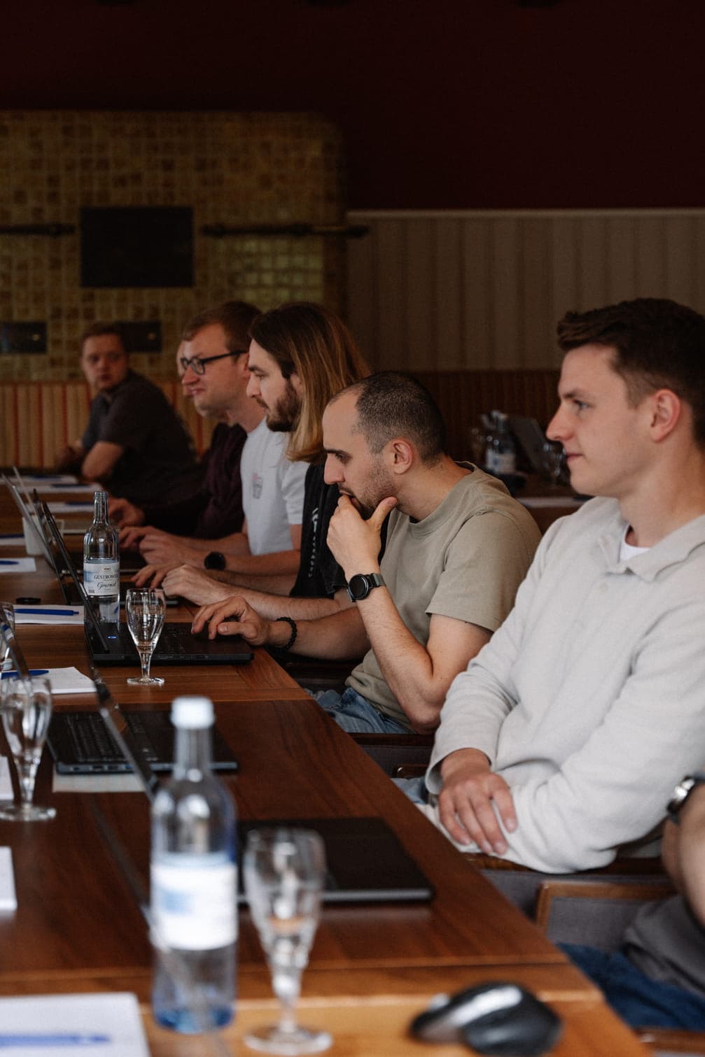 Several people seated along a long wooden table in a wood-paneled room, attending a workshop with laptops and drinks on the table.