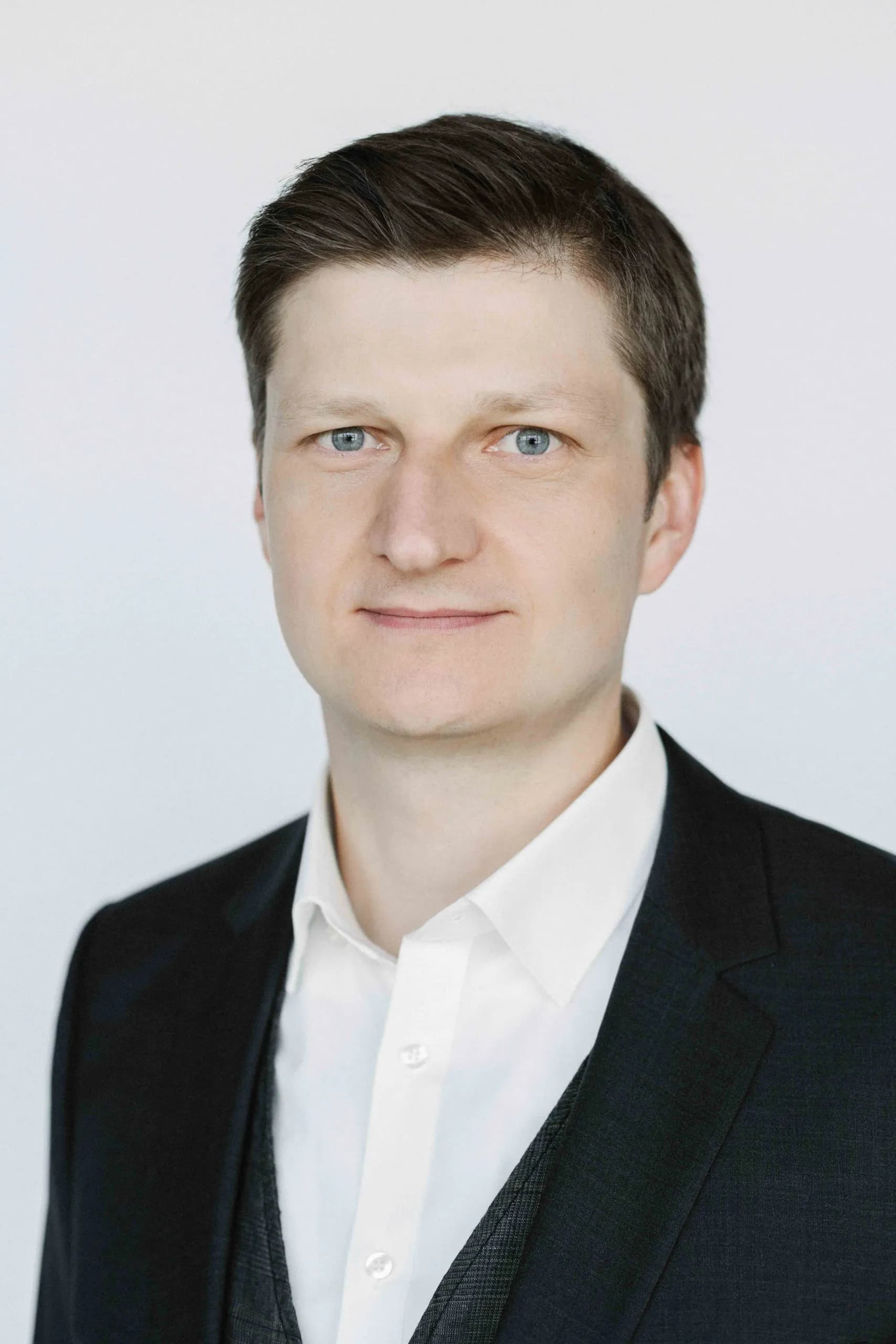 Professional headshot of Alexander Litvinenko wearing a dark blazer and open-collar white shirt against a light grey background.