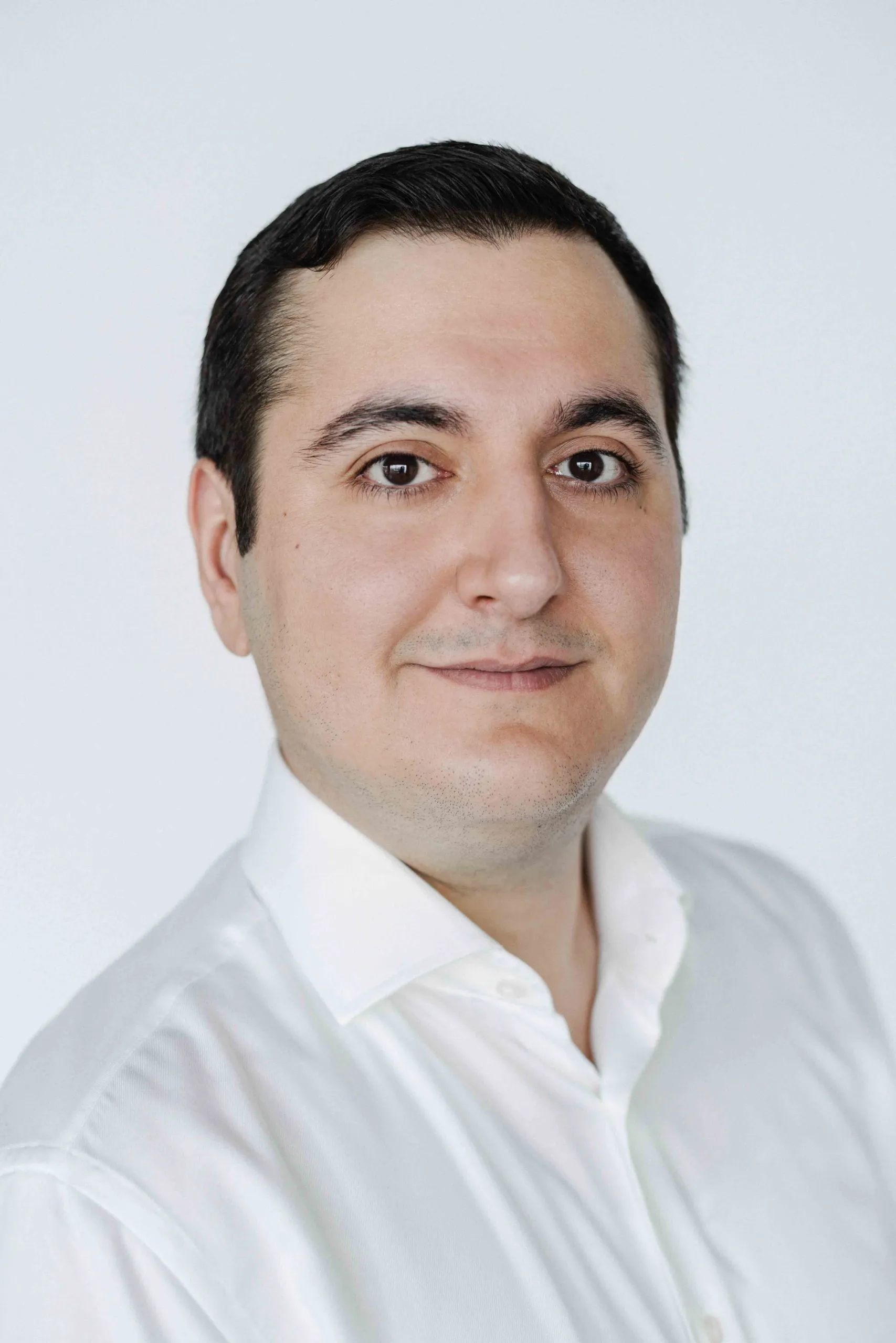 Professional headshot of a Can Aykin wearing a white collared shirt, facing the camera against a plain light grey background.
