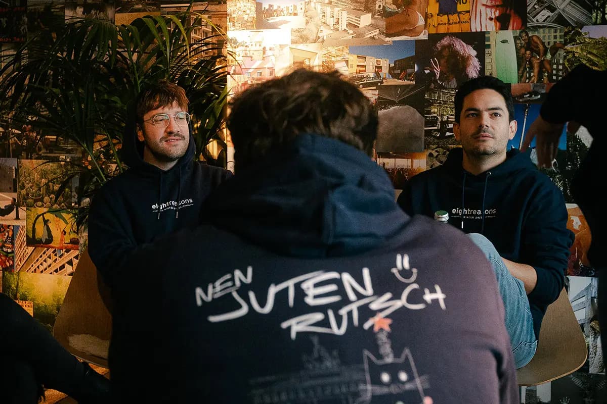 Two men in matching dark hoodies sitting and talking with a third colleague, in front of a wall covered with collage-style photos.