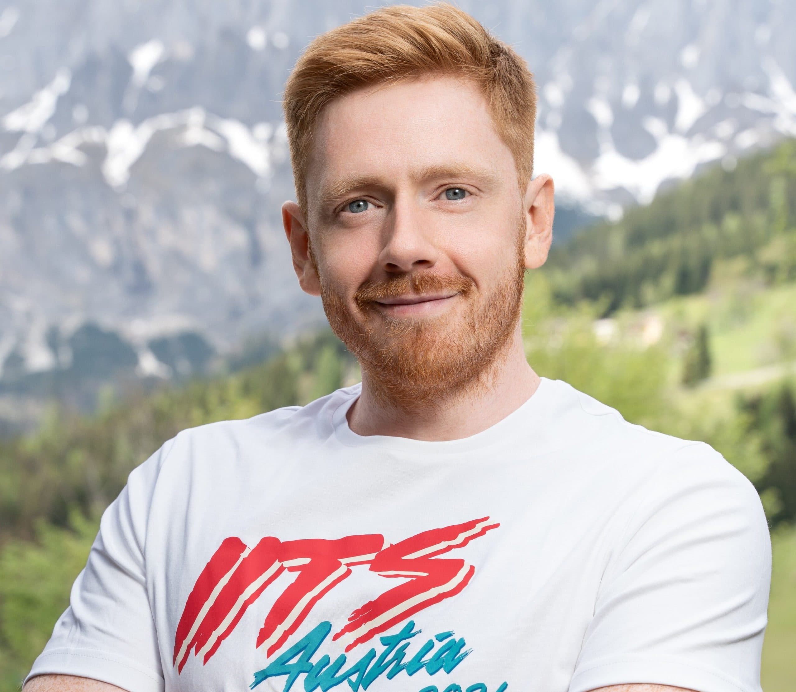 Portrait of Victor Getz, a man with short ginger hair and a matching beard, wearing a white t-shirt with an 'ITS Austria' graphic. He is standing outdoors with snow-capped mountains and green vegetation in the background.