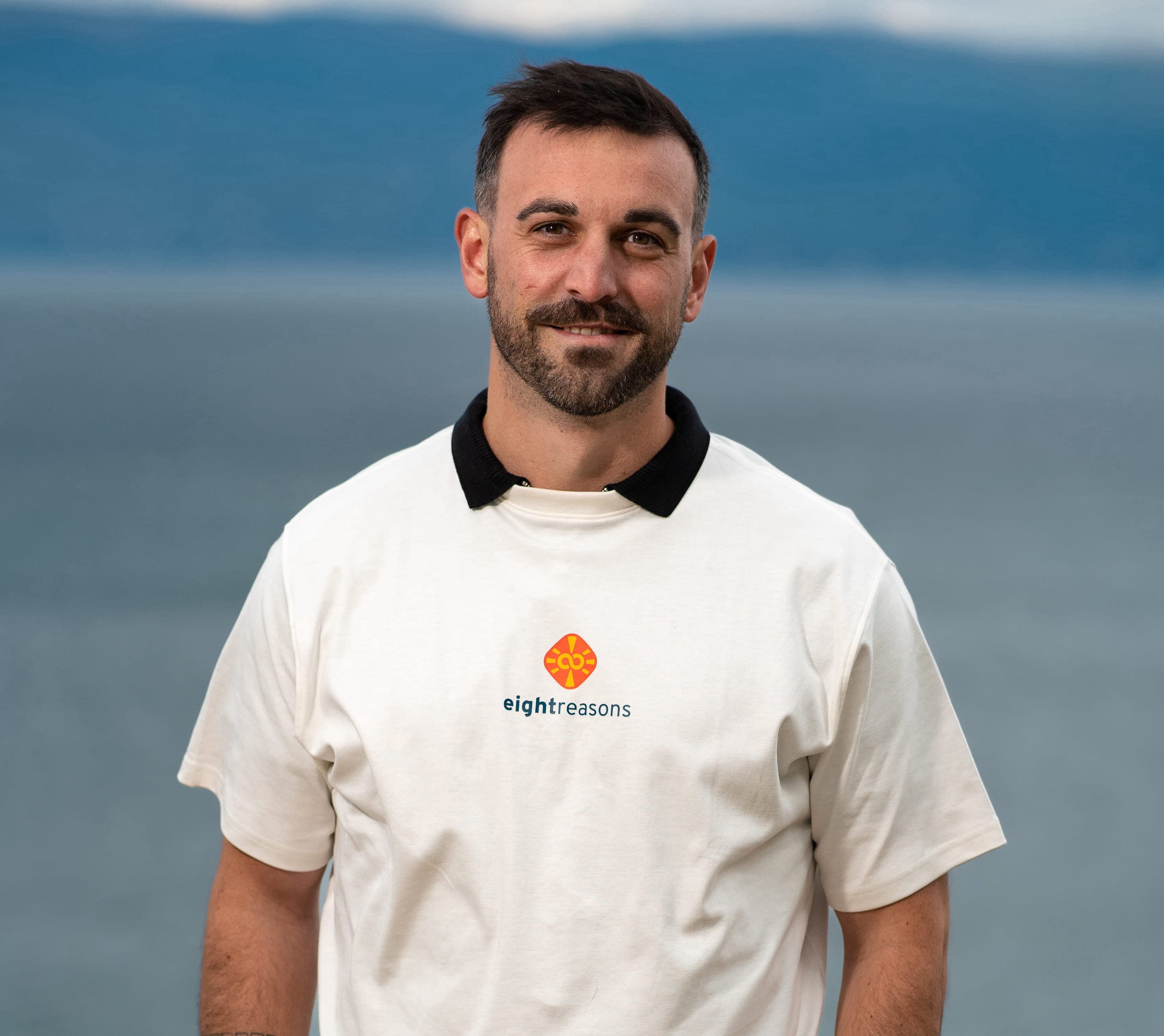 Alexander Varro, smiling with short brown hair and a beard, wearing a white polo shirt with a black collar and the eightreasons logo, against a waterside backdrop.