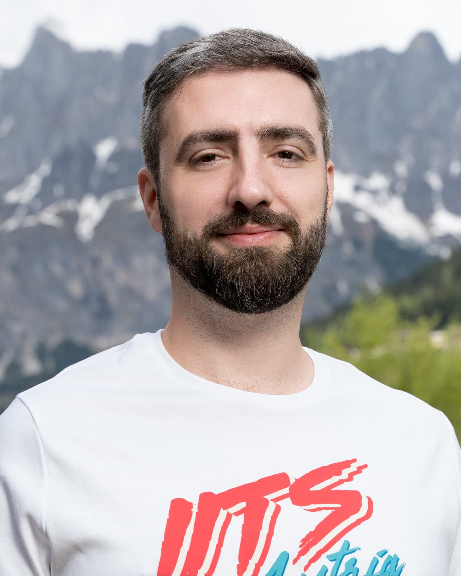 Portrait of Andreas Pongratz, a man with short, dark hair showing signs of graying and a full dark beard. He is wearing a white t-shirt with red and blue graphic lettering on the chest and smiling subtly outdoors with snow-dusted, rocky mountains and green trees in the background.