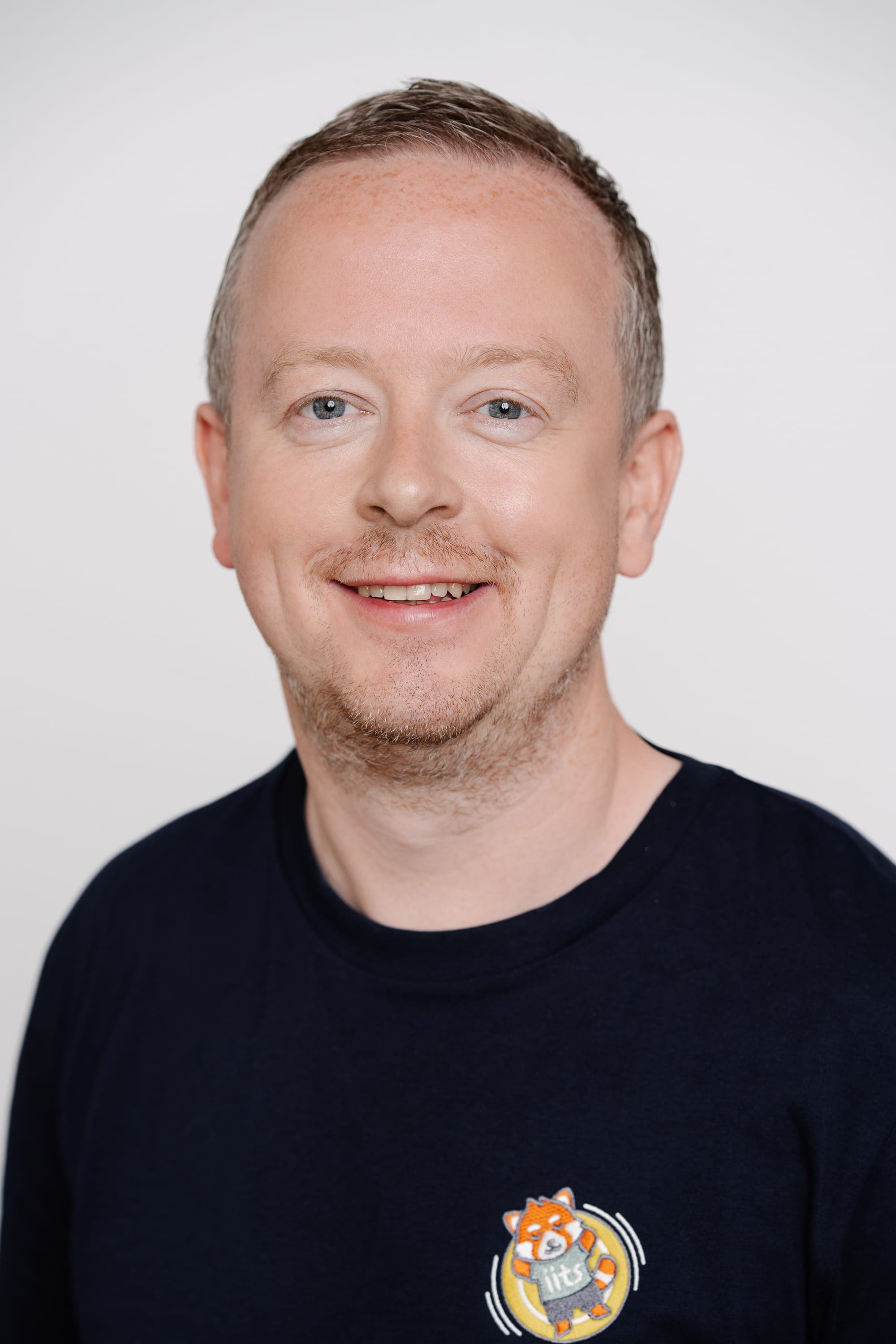 Portrait of Bernhard Kern, a man with short light brown hair, a receding hairline, and light stubble, wearing a dark blue long-sleeved shirt with a small embroidered red panda logo on the lower left. He is smiling against a plain white studio background.