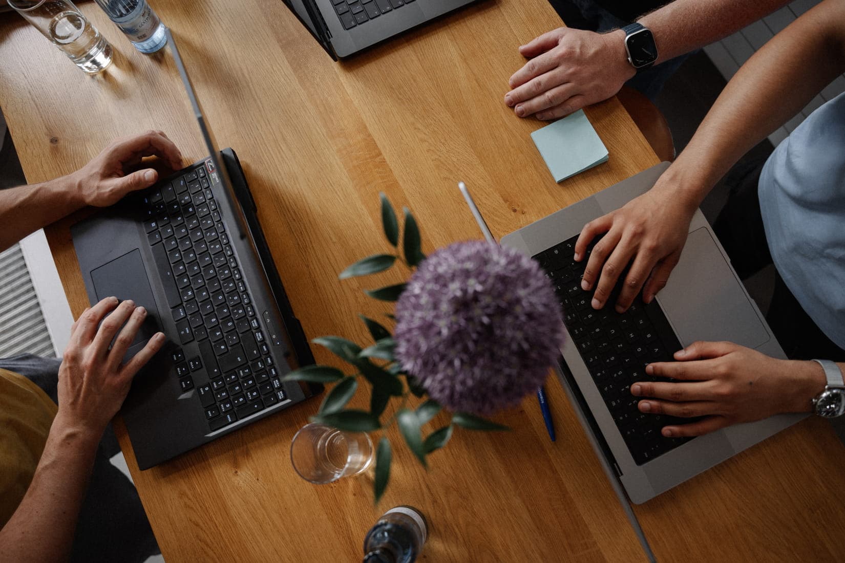 A top-down view of a collaborative workspace with laptops and hands working on a wooden table, centered around a purple flower.