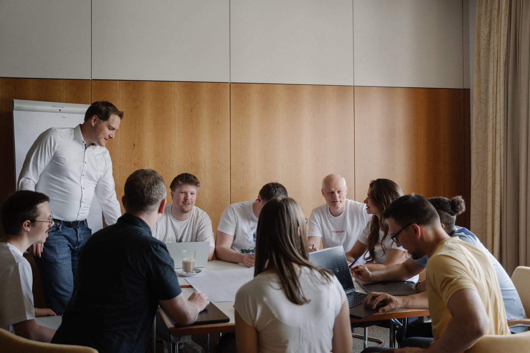 A team meeting where a standing presenter discusses documents with colleagues seated around a table with laptops.
