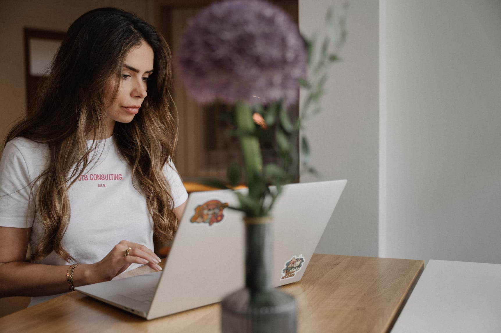 A woman with long hair works on a laptop at a wooden table, with a blurred flower vase in the foreground.