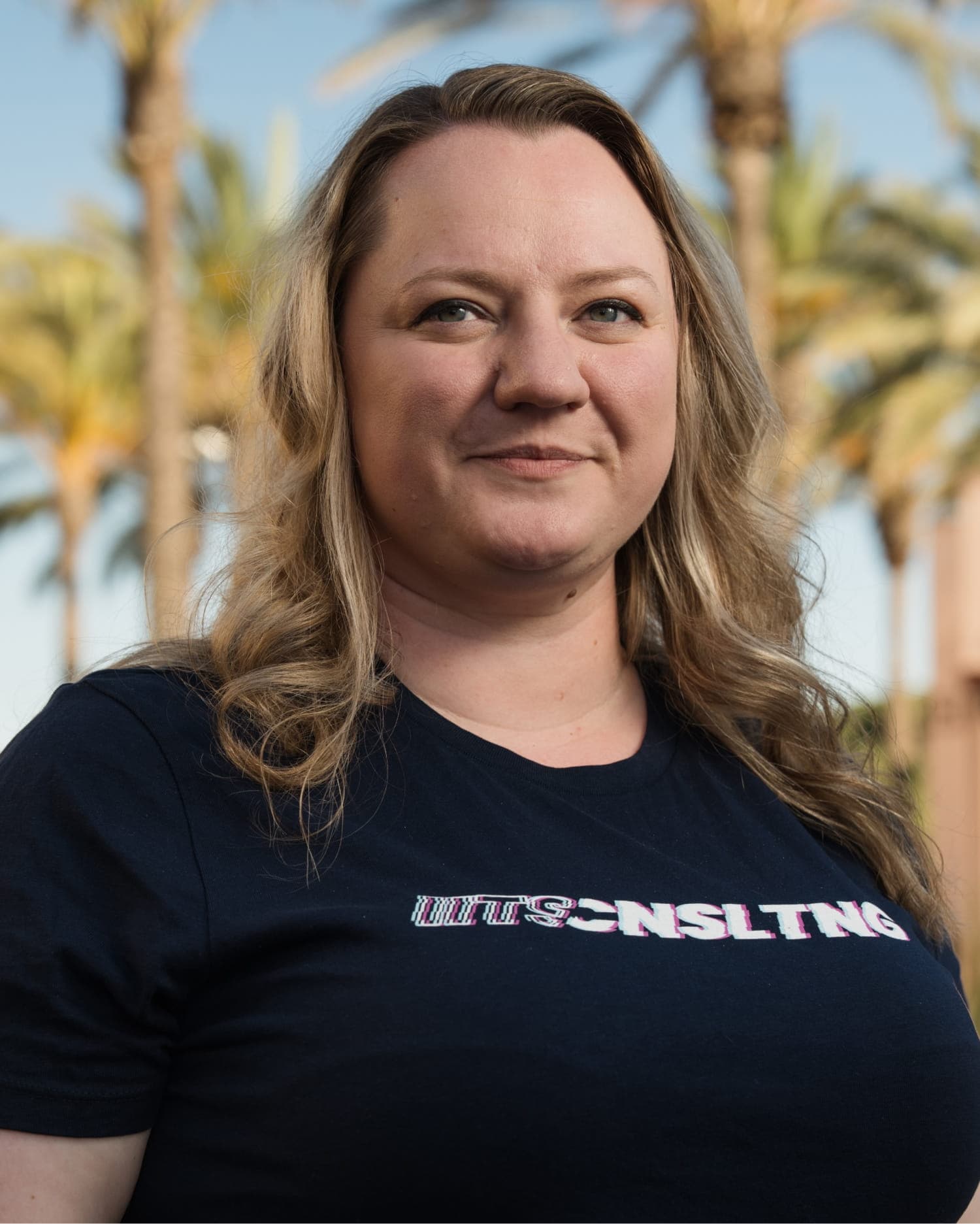 Portrait of Constanze Stohn, a woman with long, straight blonde hair, wearing a black t-shirt with a red and gold 'iits consulting' logo. She is smiling gently outdoors with green hills and mountains in the background.