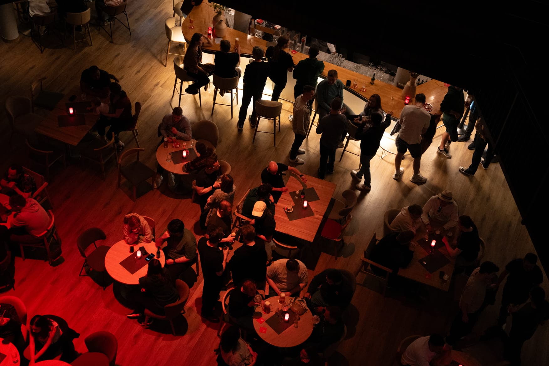 An overhead view of a busy indoor social gathering. People are seated at round tables with drinks, or standing and chatting near a long bar area.