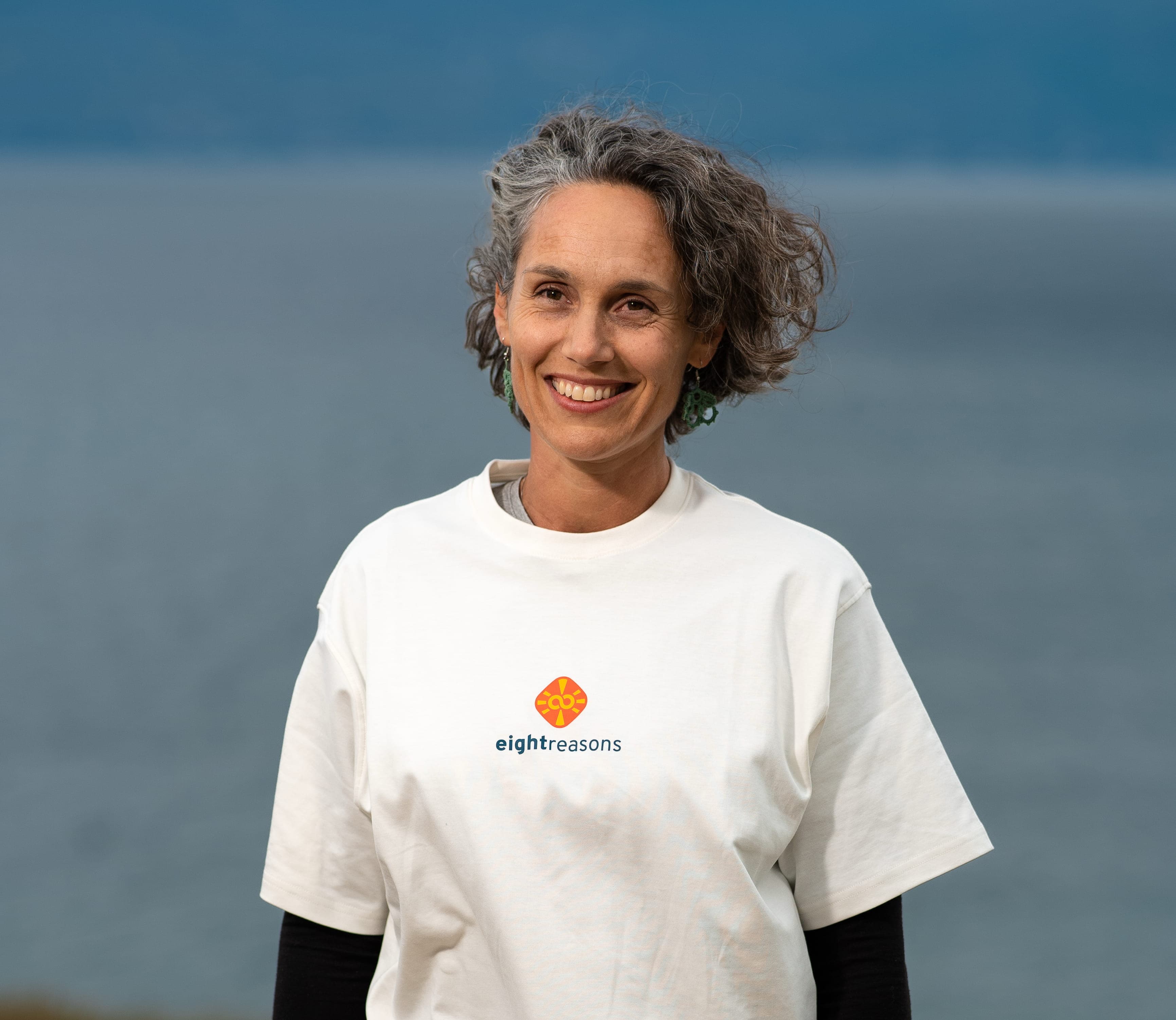 Francesca Cignola, smiling with short curly grey hair and green earrings, wearing a white eightreasons t-shirt over a black long-sleeve shirt, against a waterside backdrop