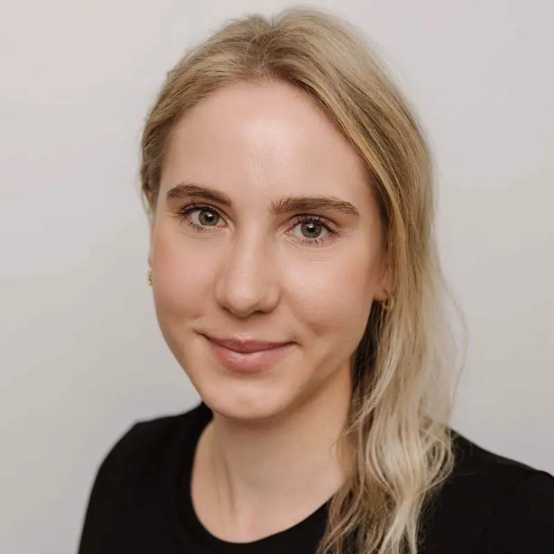 Portrait of Rafaela Heller, a young woman with long, wavy blonde hair swept over her left shoulder and green eyes. She is wearing a black crew-neck top and small gold hoop earrings, smiling gently against a plain light gray background.
