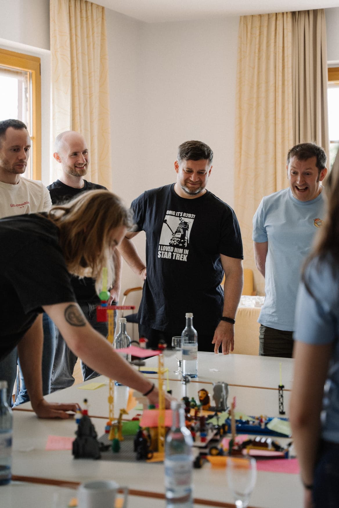 A group of colleagues gathered around a table examining colorful Lego structures, including one man wearing a black t-shirt with a Star Trek-themed graphic.