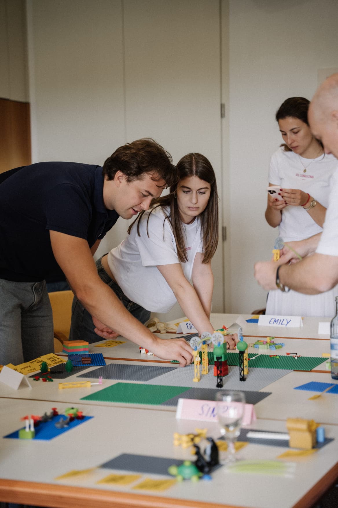A group of people working collaboratively at a table covered with LEGO Serious Play bricks and name cards. A man in a dark polo shirt and a woman in a white t-shirt are building a LEGO model together, while others in the background observe or check their phones.