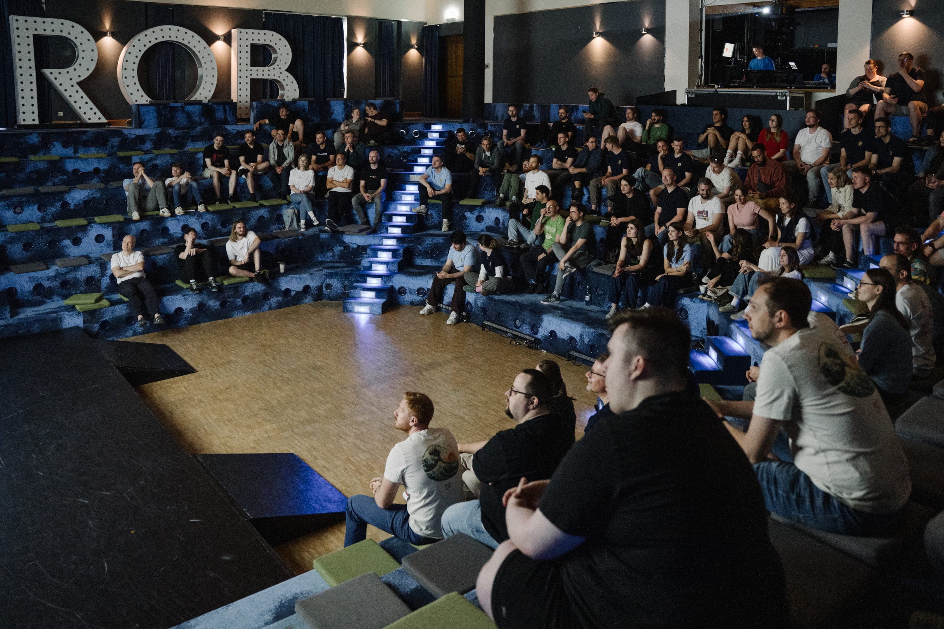 A wide-angle photograph of around 100 people seated on multi-tiered, circular blue bleachers with integrated white LED lighting along the stair treads. The bleachers surround a wooden floor area. Behind the top row, three large, illuminated marquee-style letters reading "ROB" are mounted on the dark wall, lit by warm spotlights. The audience — mostly adults dressed casually in t-shirts, hoodies, and jeans — is facing forward, watching something off-camera. In the foreground, a few people sit on the wooden floor with cushions. In the upper right background, two people can be seen in a glass-enclosed booth overlooking the room. The overall atmosphere is modern and informal.