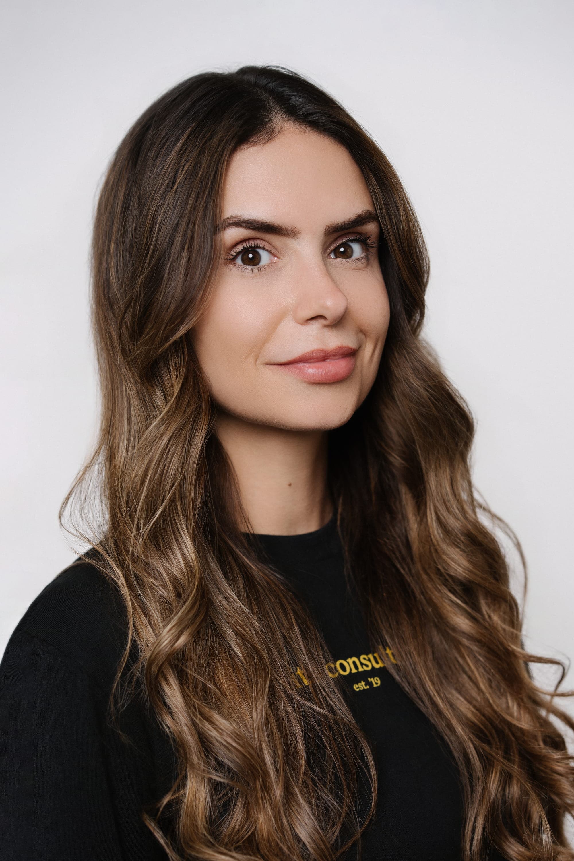 Portrait of Sarah Lössner, a young woman with long, wavy brunette hair with caramel highlights, wearing a black crewneck sweatshirt with yellow text reading 'consult est. '19'. She is smiling subtly against a plain white studio background