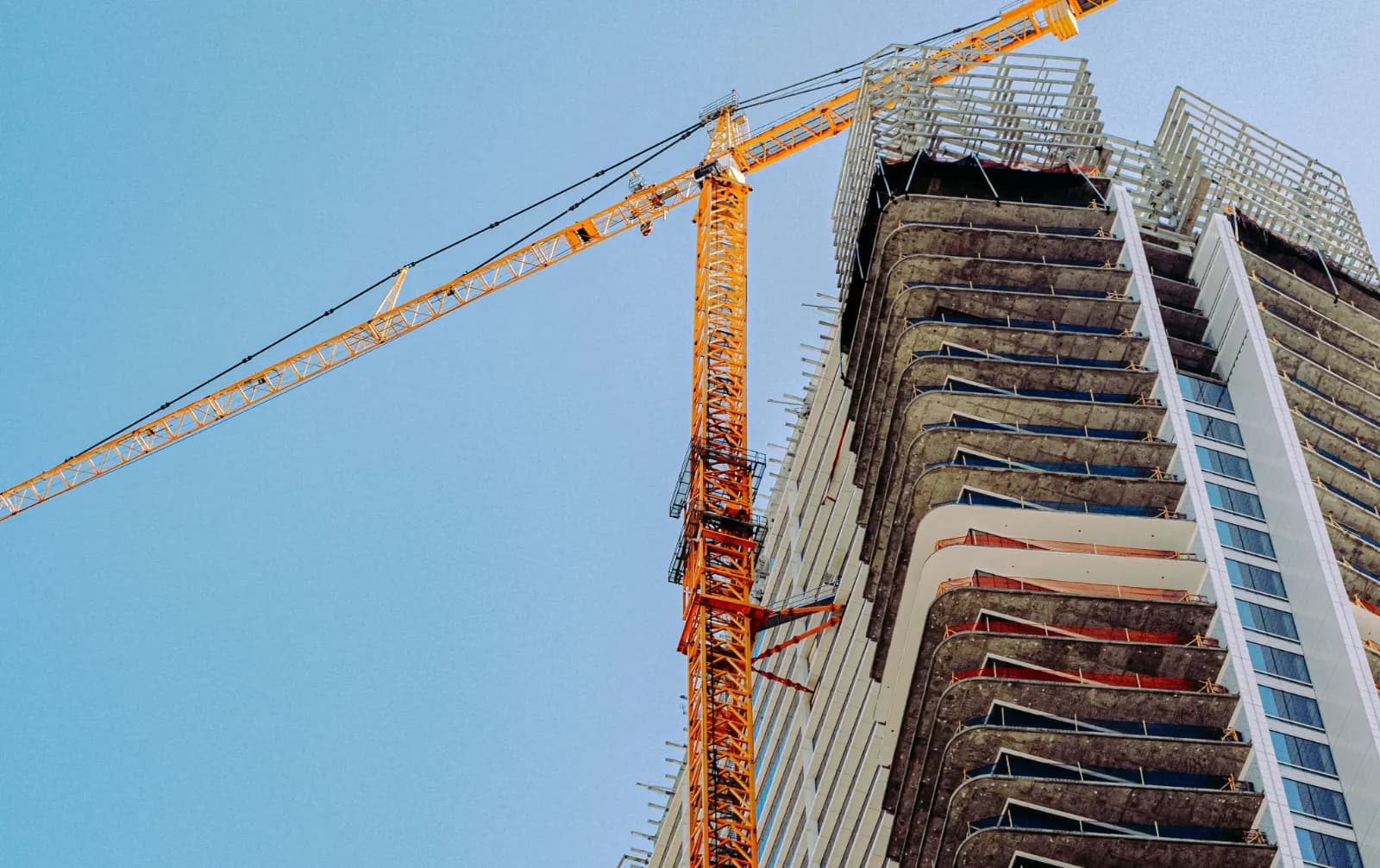 Low-angle view of a high-rise building under construction with exposed concrete floors and an orange tower crane against a clear blue sky.
