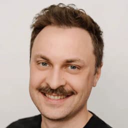 Headshot of a person with short brown hair and a mustache, smiling, wearing a black top against a light grey background.