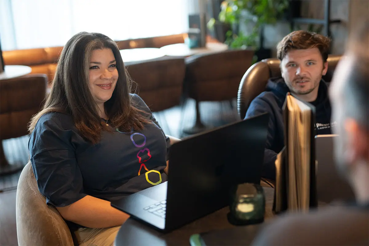 Two colleagues at a table with laptops, a woman in a branded t-shirt smiling at the camera beside a man looking her way.