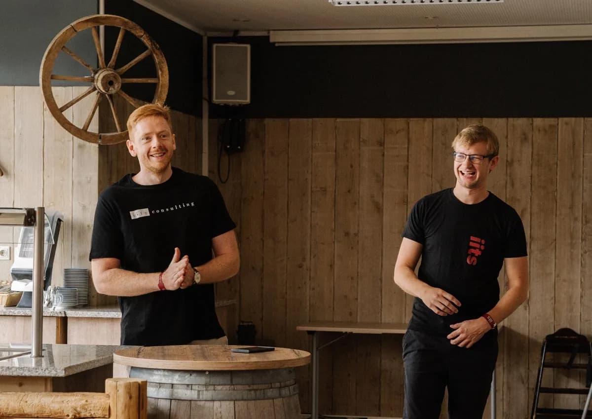 Two men standing side by side indoors against a light wood-paneled wall. On the left, Victor Getz, a man with red hair and a beard, smiles at the camera wearing a black "iits consulting" t-shirt with his hands clasped in front of him. On the right, Christoph Aufmhoff, a blonde man wearing glasses, laughs wearing a black t-shirt with a red "iits" logo. A large decorative wagon wheel hangs on the wall behind Victor.