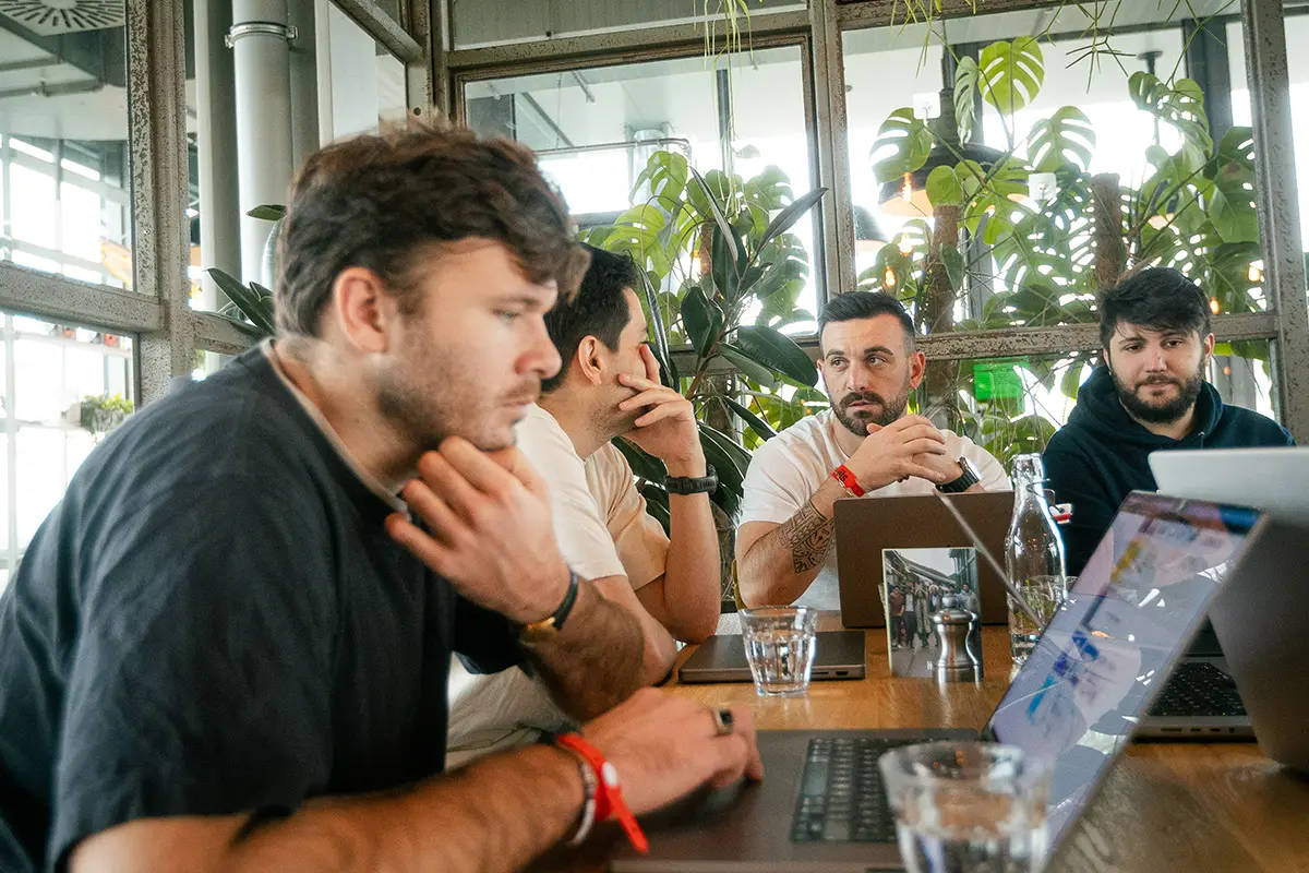 Four coworkers seated around a table with laptops and water glasses, deep in discussion surrounded by large windows and greenery.